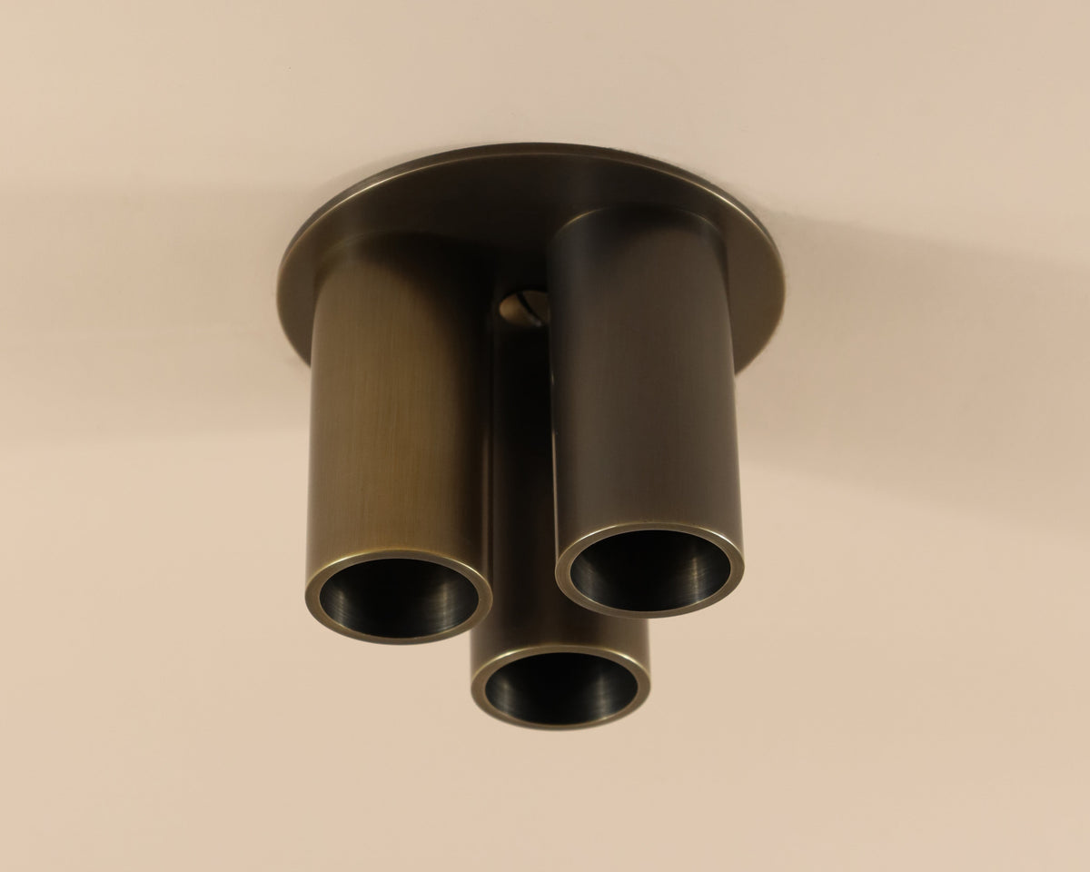 A close-up of the Long Made Co. Micro Flush Spot - Triple fixture, featuring three cylindrical metal tubes in a triangular layout on a round base, installed on a beige ceiling.