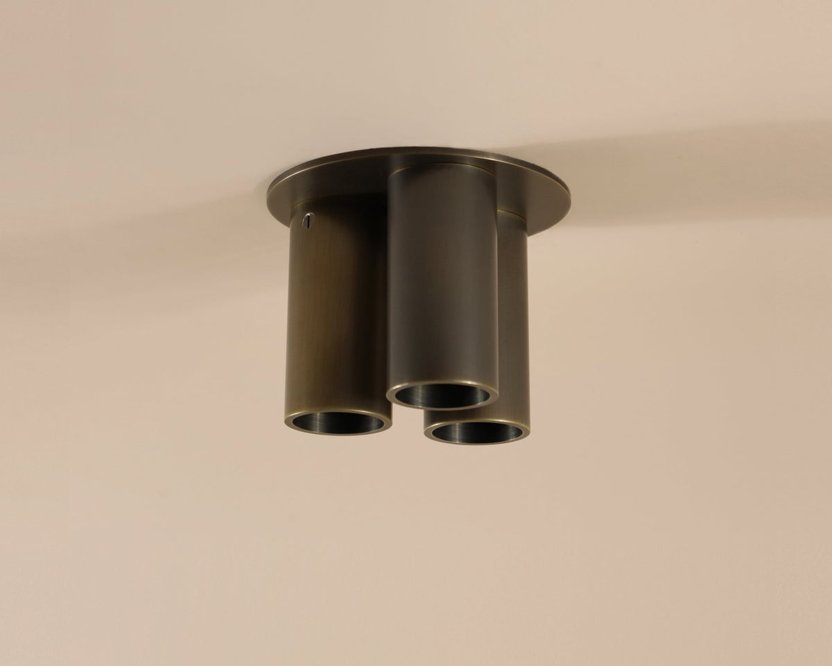 The Long Made Co. Micro Flush Spot - Triple is a handmade lighting fixture with three cylindrical, dark metallic tubes in a triangular pattern on a round base, creating an elegant ultra flush mount look against a beige ceiling.