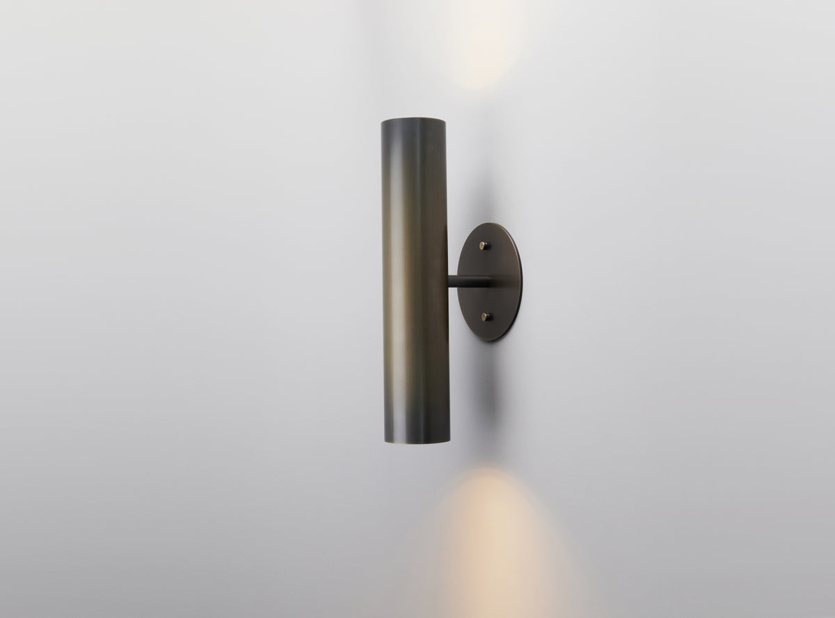 The Brass Cylinder Up-Down Sconce by Long Made Co. features a sleek cylindrical design with a black finish, casting light both upward and downward against a plain white wall to evoke the timeless elegance of mid-century modern lighting.