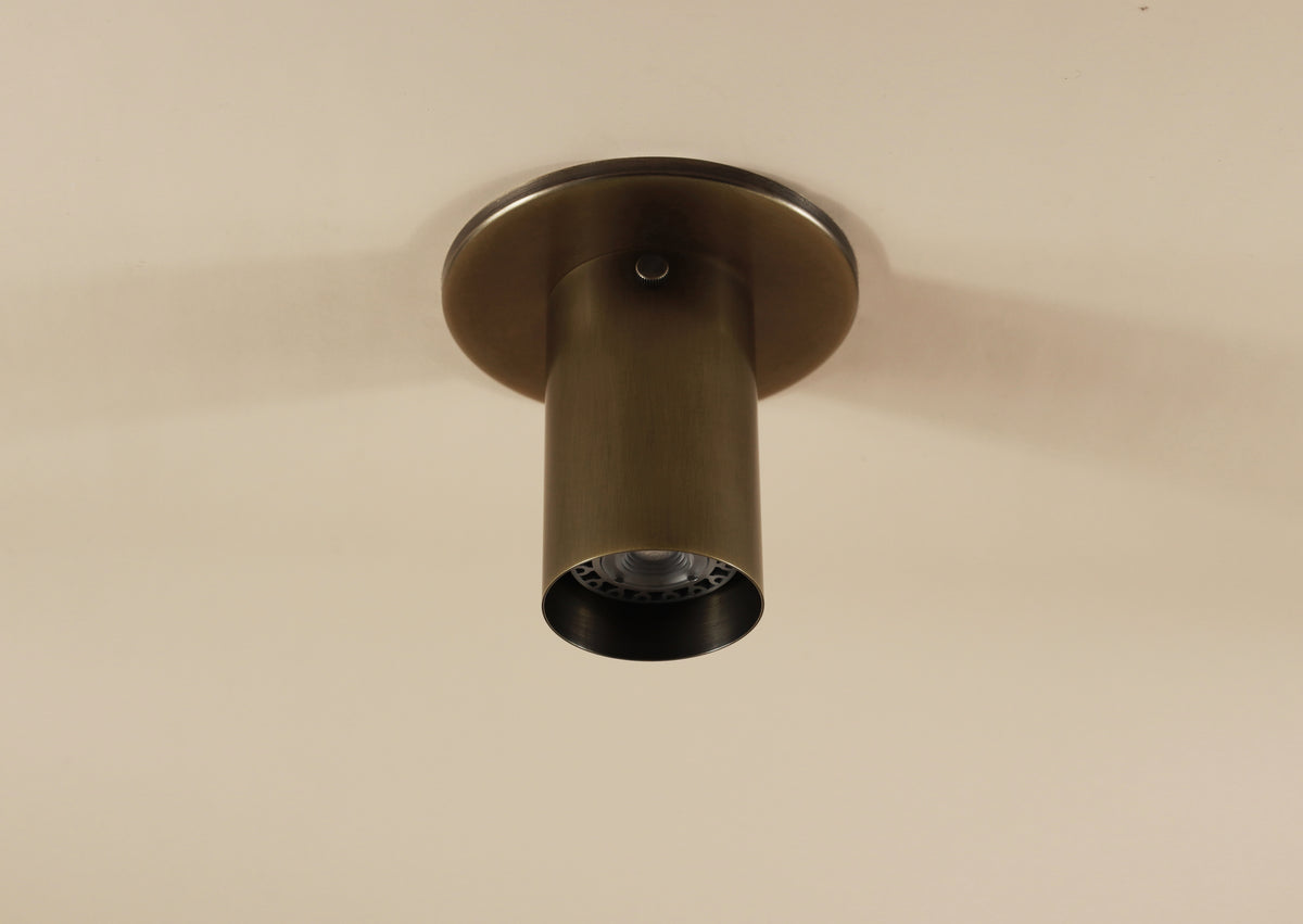 The Brass Cylinder Flush Spot by Long Made Co. is a solid brass ceiling fixture with a simple, modern design and partially recessed bulb, mounted on a flat beige ceiling, inspired by classic mid-century modern style.