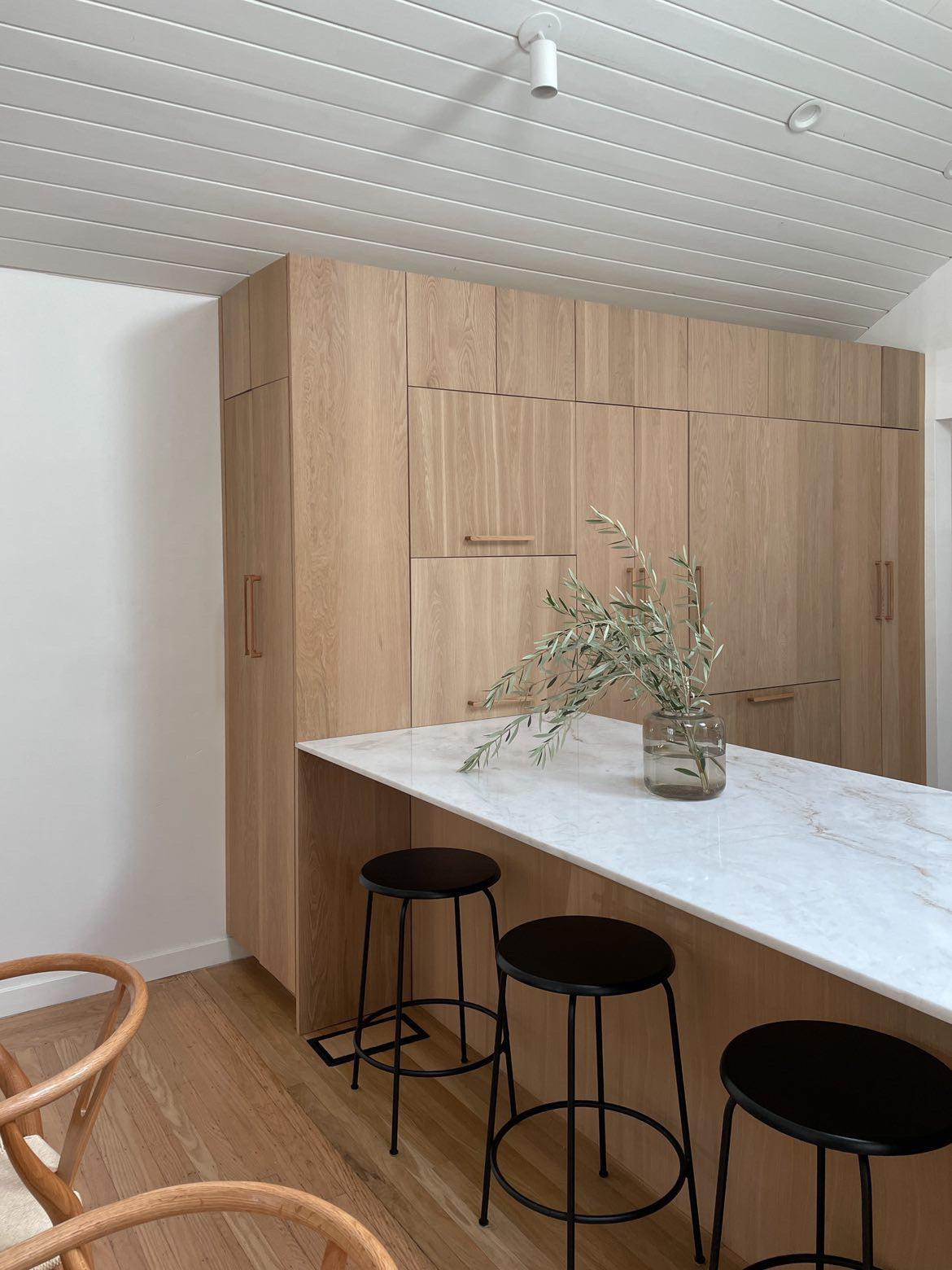 A modern kitchen with light wood cabinets, a white marble island, black metal stools, and a glass vase with green branches is enhanced by Cove Adjustable Spot lights from Long Made Co., which highlight the clean, minimalistic design.