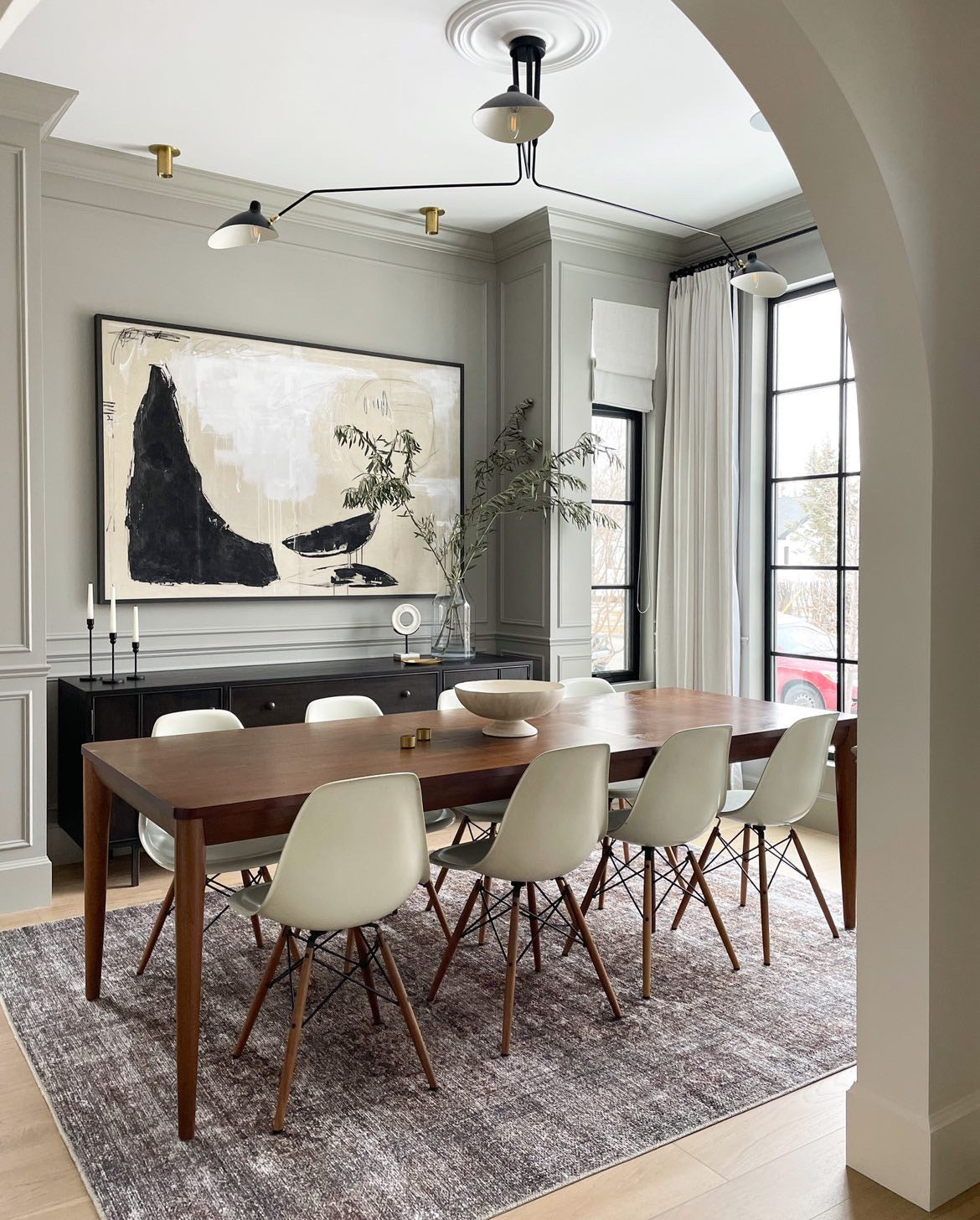 A modern dining room featuring a wooden table, six white chairs, a gray rug, and large abstract wall art. Black-framed windows, light gray walls, and Long Made Co.'s Brass Cylinder Flush Spot lighting create an elegant atmosphere.
