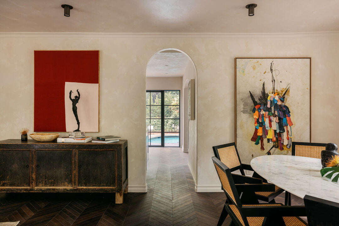 A stylish dining room features a marble table, black chairs, eclectic art on cream walls, a rustic sideboard, and the Brass Cylinder Flush Spot by Long Made Co. An arched doorway opens to a bright room with large windows and pool views.