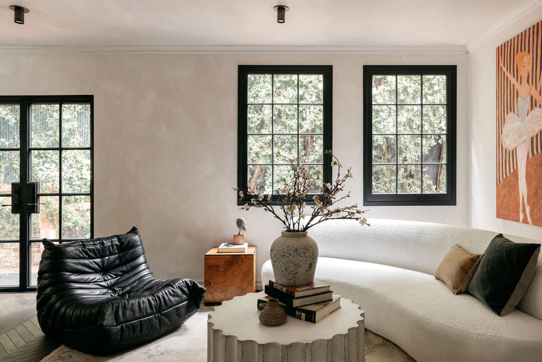 A modern living room with large windows, a white curved sofa, and a black lounge chair features the Brass Cylinder Flush Spot by Long Made Co. A round coffee table, books, a vase, and a side table with lamp complete the neutral-toned space.