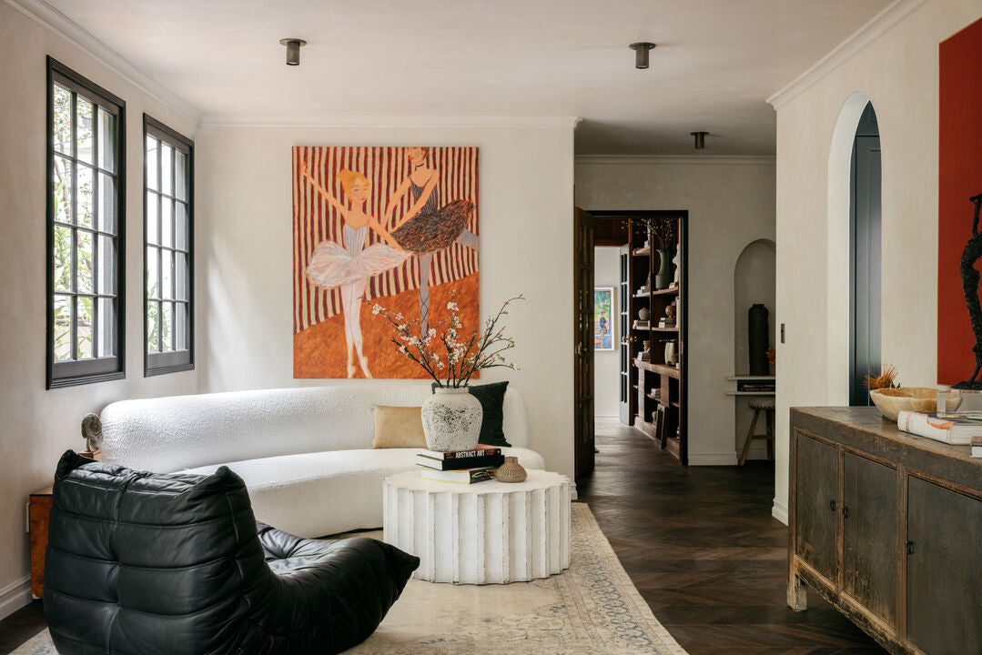 A modern living room with a curved white sofa, black leather lounge chair, round white coffee table, and large ballerina painting; natural light pours in under the Long Made Co. Brass Cylinder Flush Spot.
