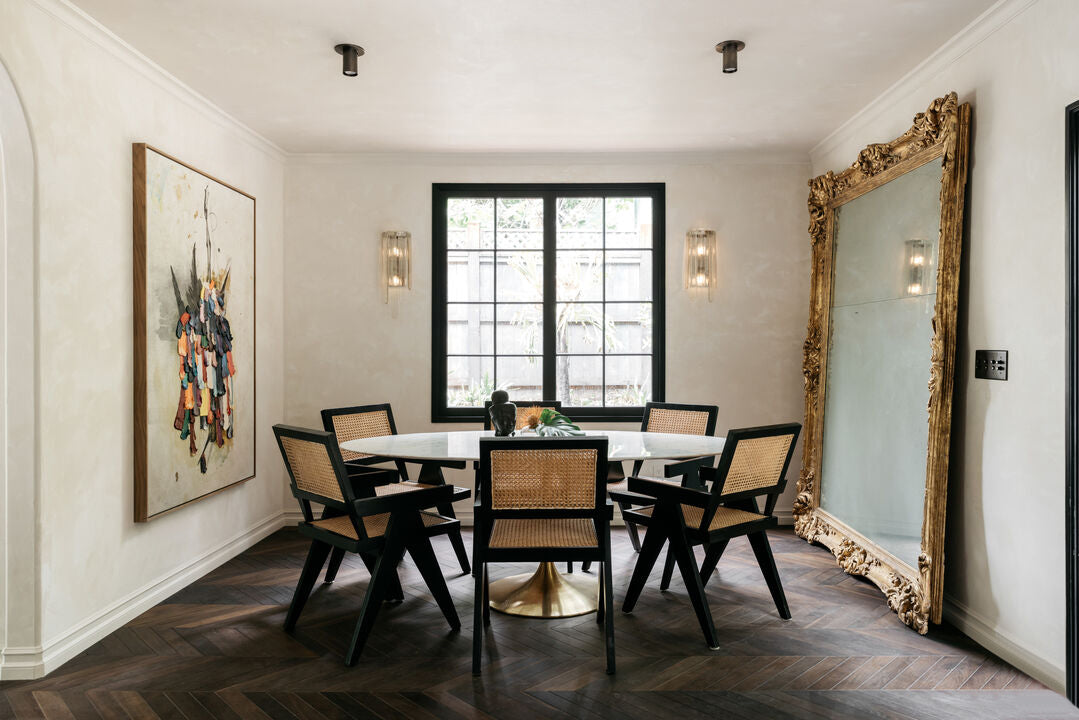 A stylish dining room features a round white table, six black chairs with cane seats, a large gold-framed mirror, and colorful art. The Brass Cylinder Flush Spot by Long Made Co. adds mid-century flair above the dark herringbone wood floor.