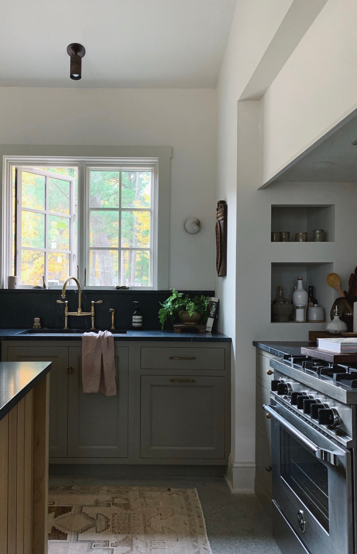 A bright kitchen with gray cabinets, brass faucet, potted herbs, and open shelves is illuminated by Cove Adjustable Spot lights from Long Made Co. Sunlight streams in above the sink, highlighting jars and a black stove on the right in this modern space.