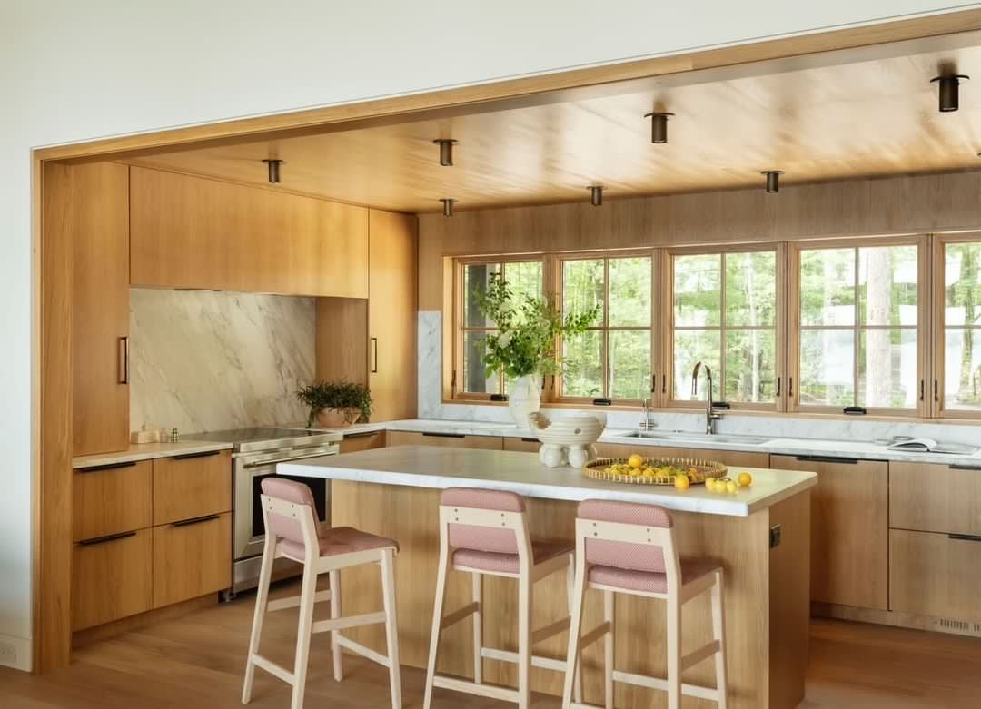 Modern kitchen with wooden cabinets, marble countertops, and an island with three pink barstools. Large windows bring in light, while the Brass Cylinder Flush Spot by Long Made Co. adds mid-century modern style above lemons and a fruit bowl on the island.