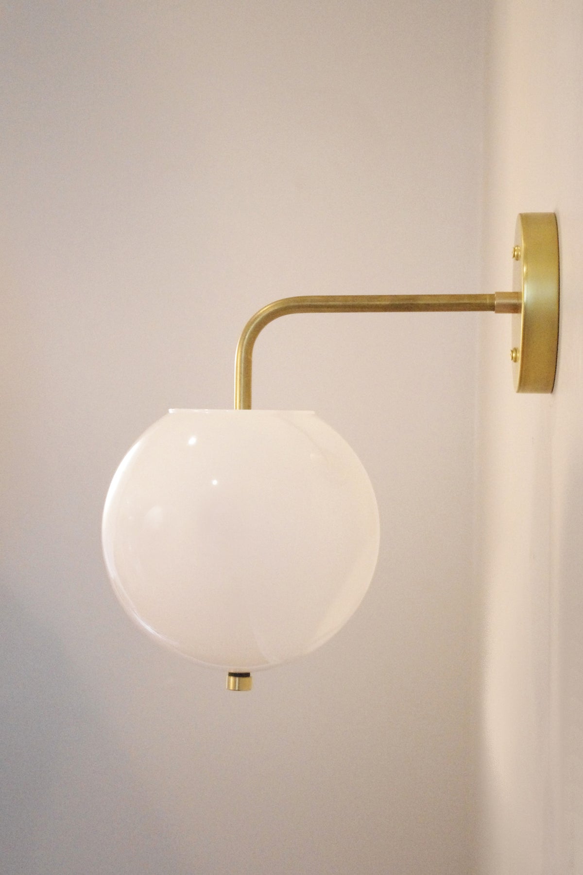 The Long Made Co. Wade Wall 8" Globe is a modern wall sconce featuring a handmade brass arm and a round white glass globe shade, shown mounted on a light-colored wall.