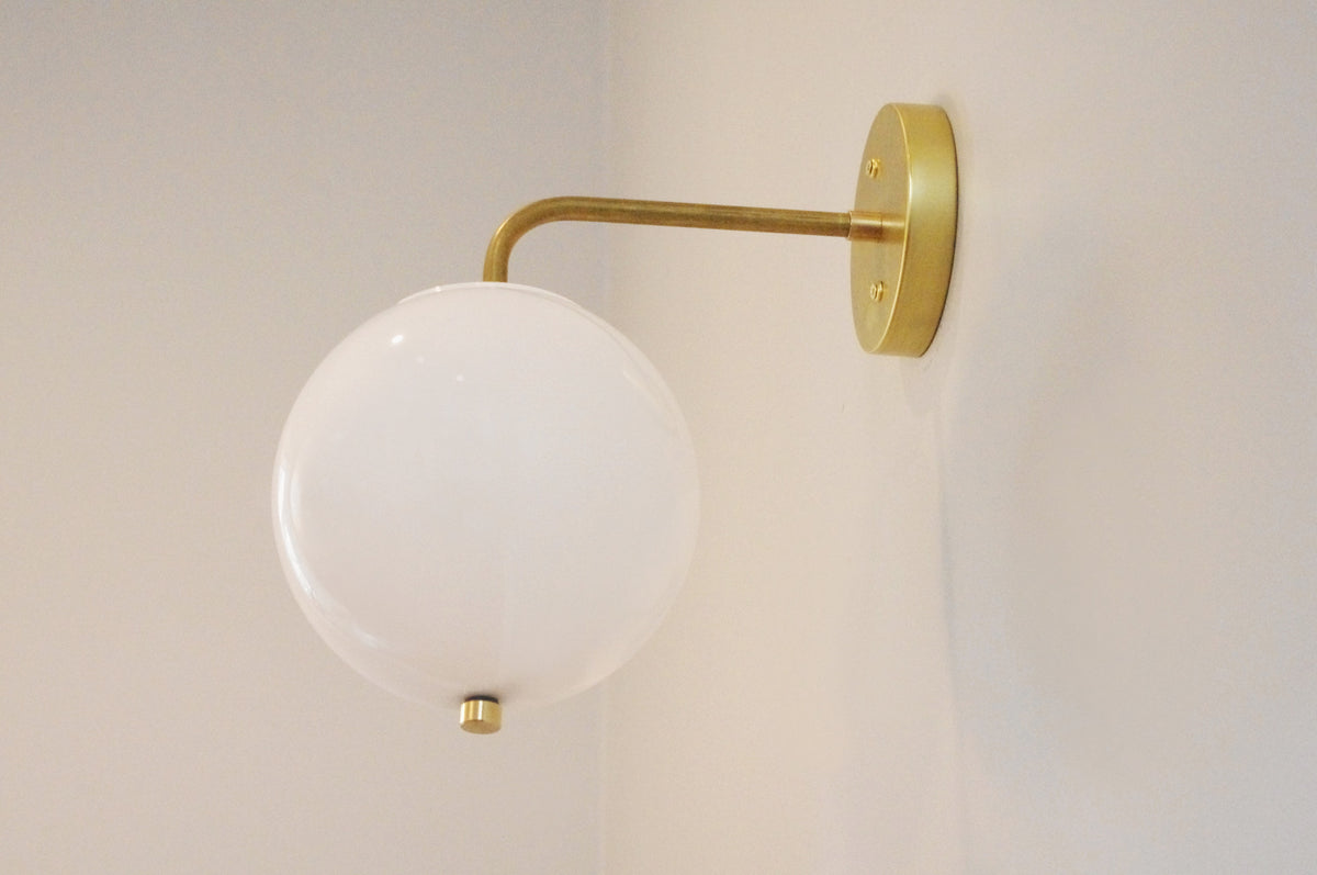 The Wade Wall 8" Globe by Long Made Co. is a modern wall sconce with a handmade brass arm and an acrylic globe shade, mounted on a light-colored wall.