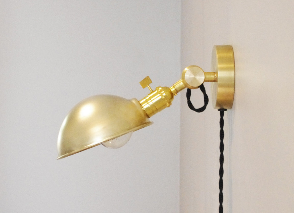 The Task Loop Wall Lamp - Parabolic Shade by Long Made Co., featuring a gold-colored finish, adjustable arm, dome-shaped shade, and black twisted cord, is mounted on a white wall to serve as a stylish handmade brass lighting solution—perfect as a bedside reading light.