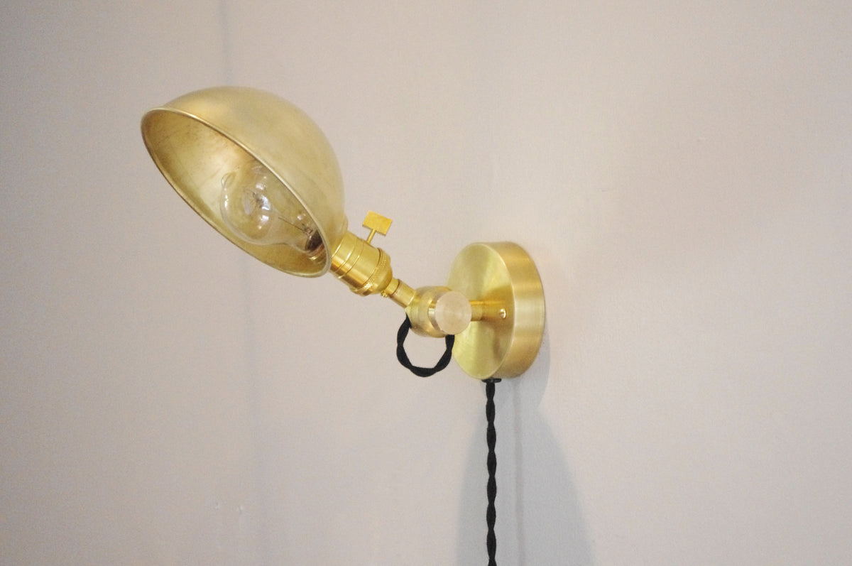 Introducing the Task Loop Wall Lamp - Parabolic Shade by Long Made Co.—a handcrafted brass wall-mounted fixture featuring a round base and an adjustable arm. Perfect as a plug-in bedside reading light, its sleek brass design stands out against a light-colored wall, while an elegant black twisted cord hangs from the lamp.
