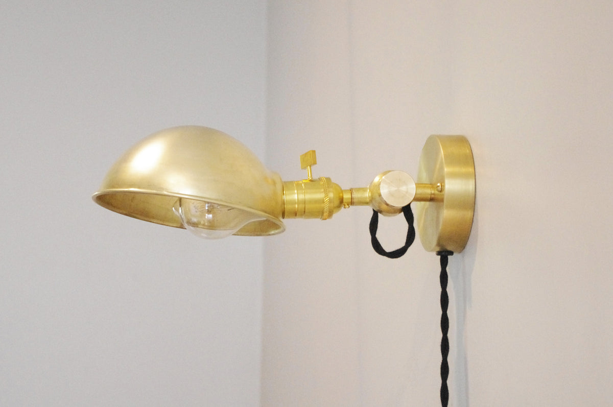 Introducing the Long Made Co. Task Loop Wall Lamp - Parabolic Shade: a handmade brass wall-mounted sconce featuring a distinctive dome-shaped (parabolic) shade, exposed bulb, round base, and twisted black cord. Designed as an easy-to-install plug-in wall lamp, it attaches effortlessly to any white wall—making it an ideal choice for a stylish and functional bedside reading light.