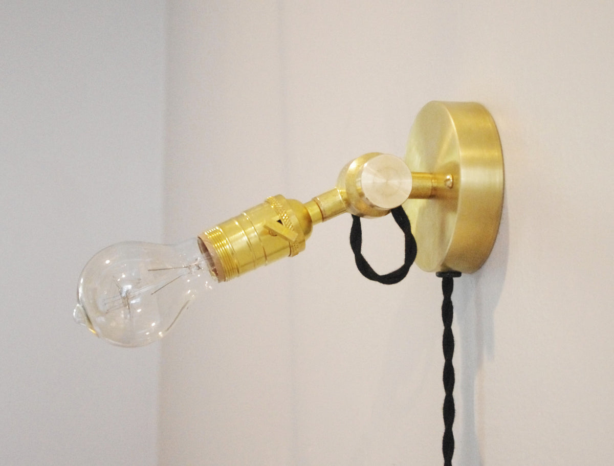 The Task Loop Wall Lamp by Long Made Co. is a handmade brass lighting fixture designed for wall mounting, featuring an exposed bulb with a round base and a black twisted cord draping elegantly against a white wall. This plug-in wall light offers both stylish appeal and convenient installation for any space.