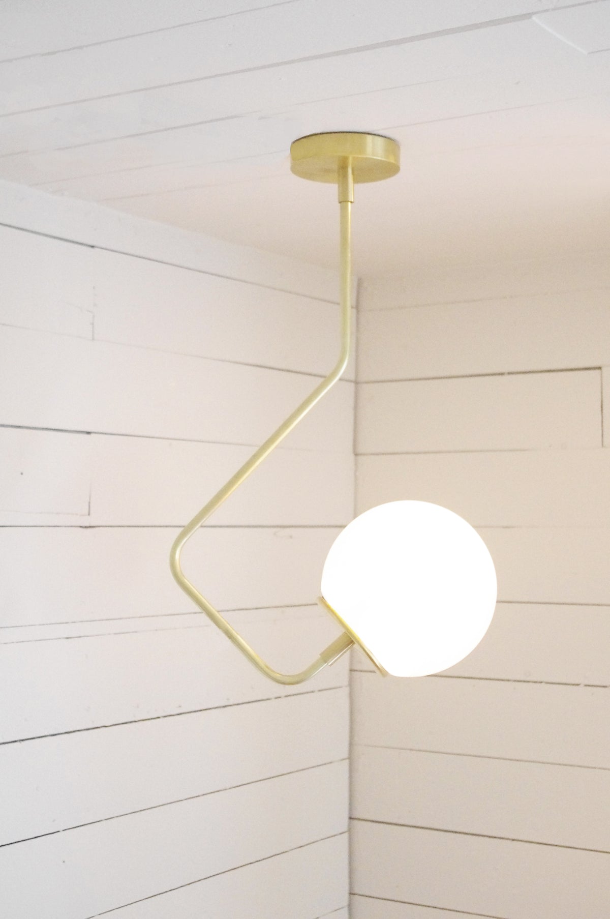 The Sphere Solo Bent Arm by Long Made Co. features a geometric, angular brass arm and a hand-blown glass globe, creating a modern custom ceiling light ideal for mounting on white shiplap ceilings or walls.