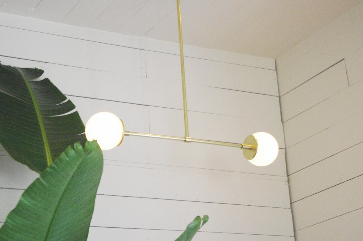 The Long Made Co. Globe Double Chandelier, a modern solid brass fixture with two round white bulbs, hangs from the ceiling against white shiplap walls. Large green tropical leaves appear in the lower left corner.