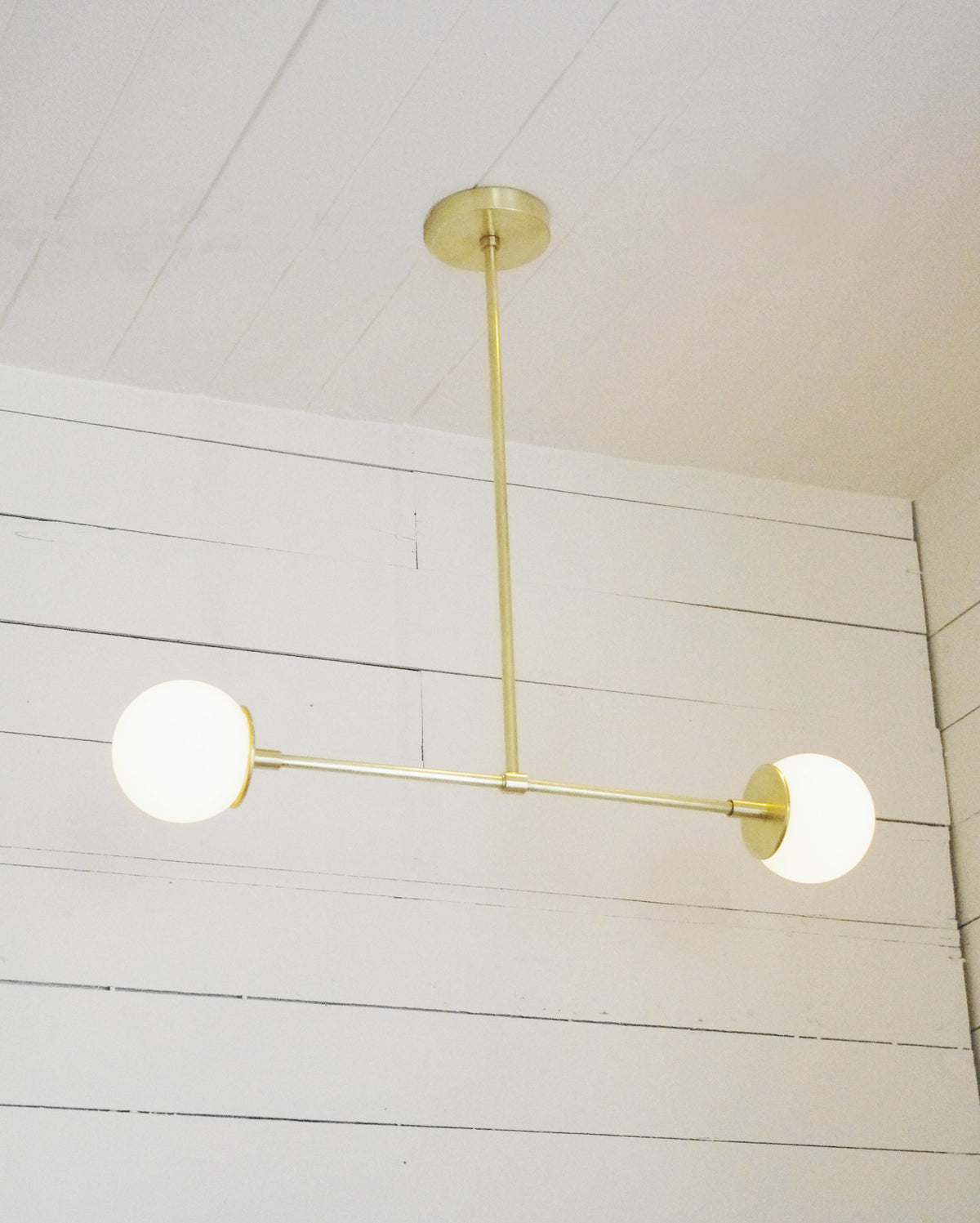 The Globe Double Chandelier by Long Made Co. features a solid brass fixture with two round white globes, suspended from a white paneled ceiling.