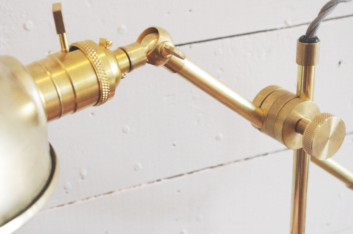 Close-up of the Task Articulating Table Lamp by Long Made Co., featuring a brass finish, adjustable arm, and knurled knobs, set against a white wooden wall with visible screw holes.
