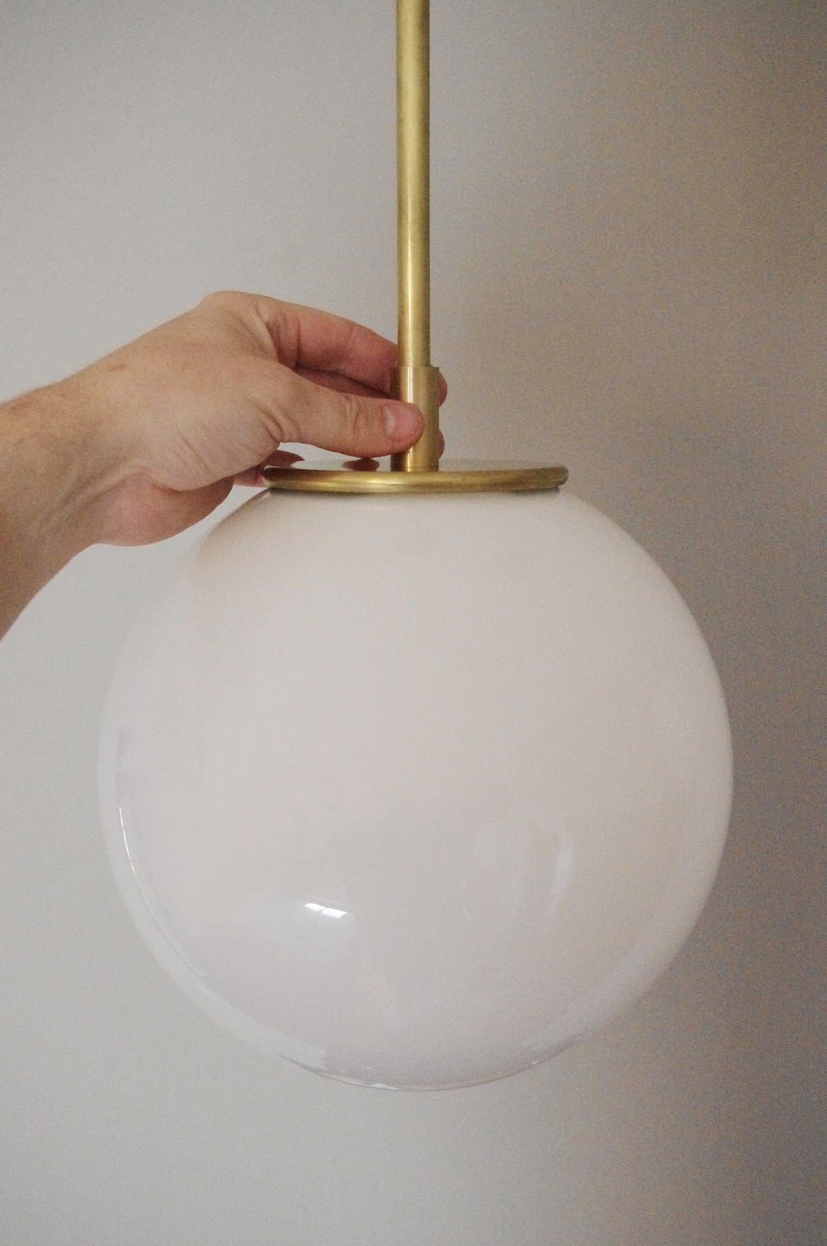 A hand holds the Long Made Co. Sphere Solo Drop - 10" pendant light, crafted from solid brass with a modern spherical opal glass shade, against a plain, light-colored wall.