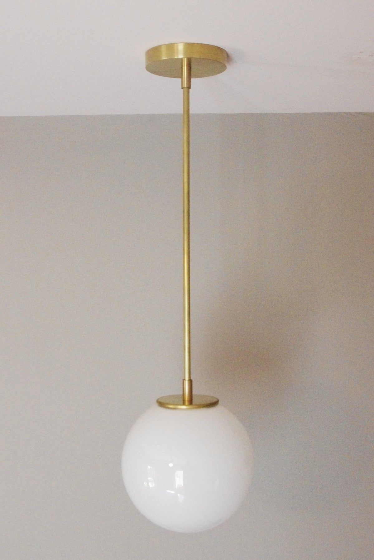 The Sphere Solo Drop - 10" by Long Made Co. is a modern solid brass pendant with a brass rod and fittings, featuring a round opal glass shade and showcasing elegant handmade craftsmanship against a light gray wall.