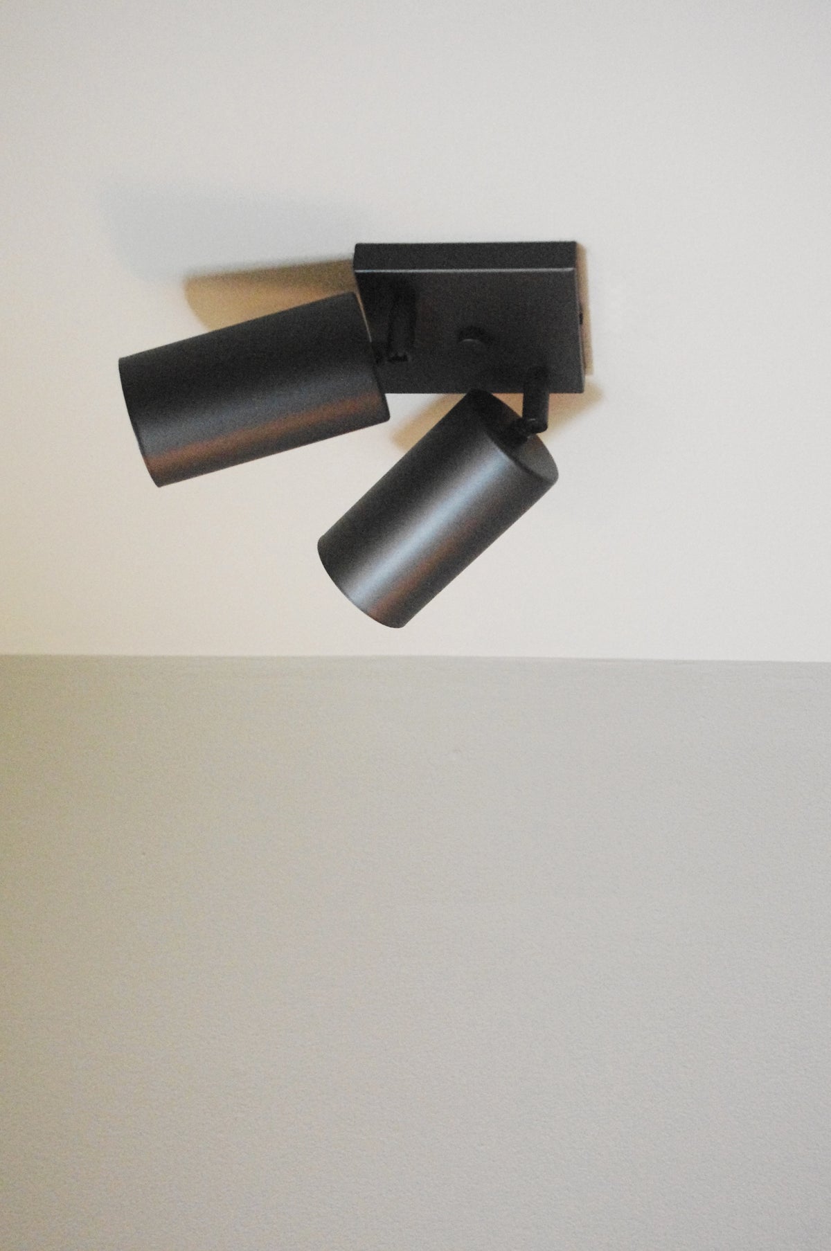 The Cove Double Spot by Long Made Co., a black ceiling-mounted downlight with two adjustable cylindrical lights, is installed on a white ceiling above a light gray wall.
