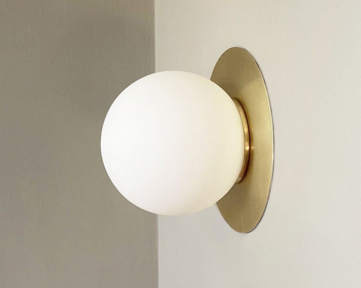 A wall-mounted light fixture with a gold-colored backplate and a white spherical opal shade, viewed from a side angle against a neutral background.