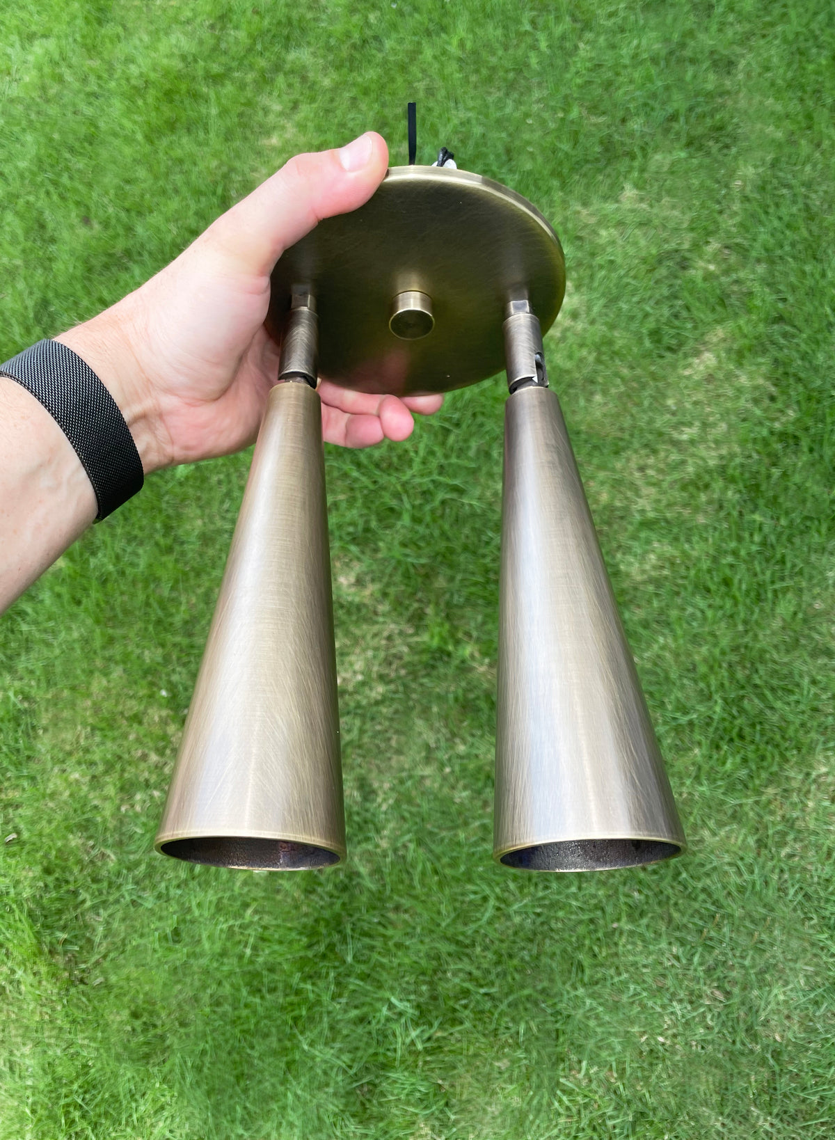 A hand holds the Pine Double Spot by Long Made Co., a handmade in Texas, brass-colored fixture with adjustable spot heads on a circular base, shown outdoors over green grass and both heads facing downward.