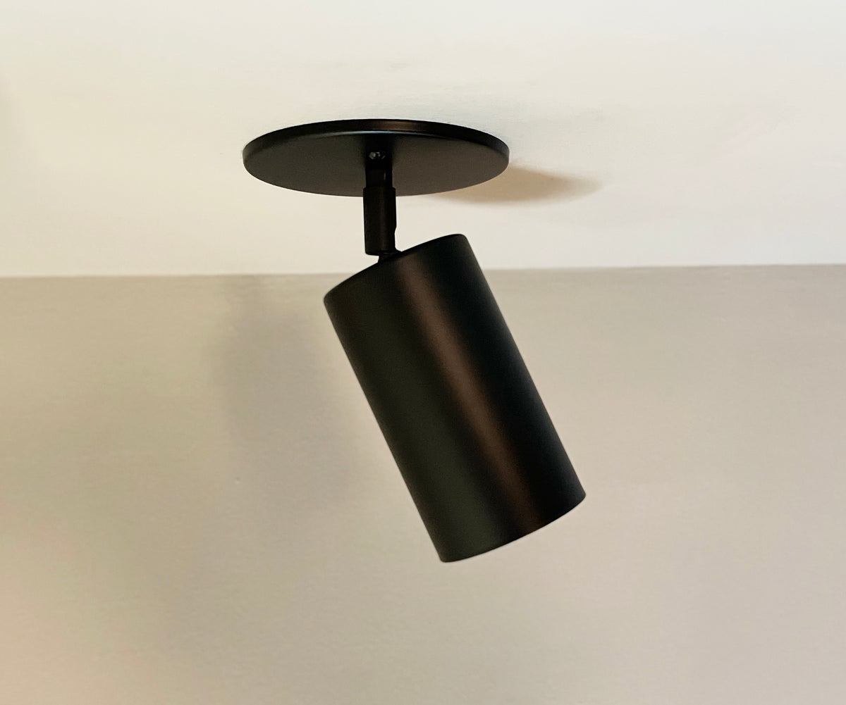 The Cove Wall Lamp by Long Made Co. is a black, cylindrical ceiling spotlight with a minimalist design, mounted on a round base and angled downward against a white ceiling and light-colored wall.