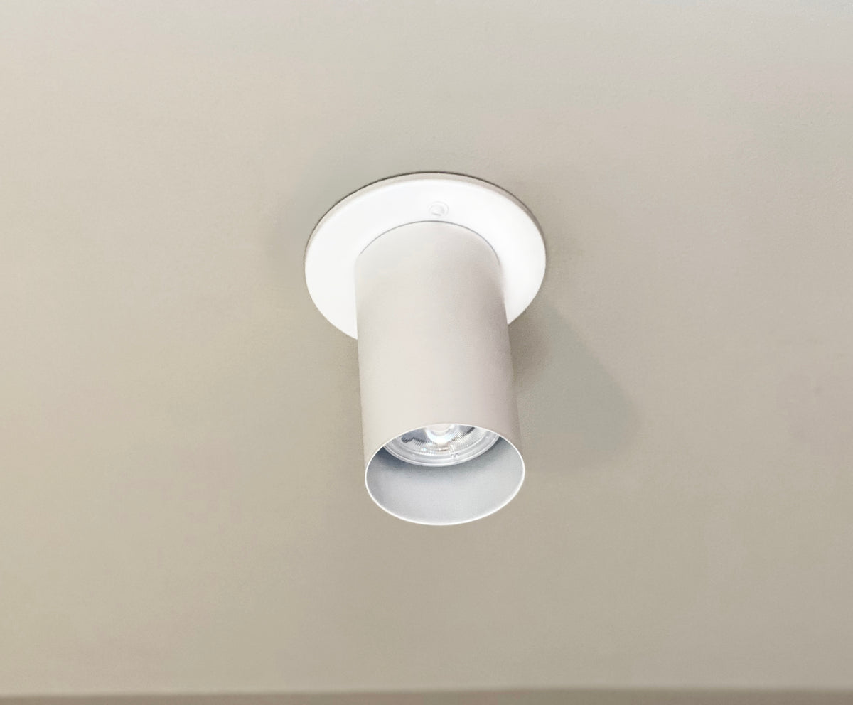 The Cove Flush Spot by Long Made Co. is a white cylindrical ceiling light with a minimalist design, exposing the bulb for modern appeal, and is flush-mounted on a smooth, light-colored ceiling.