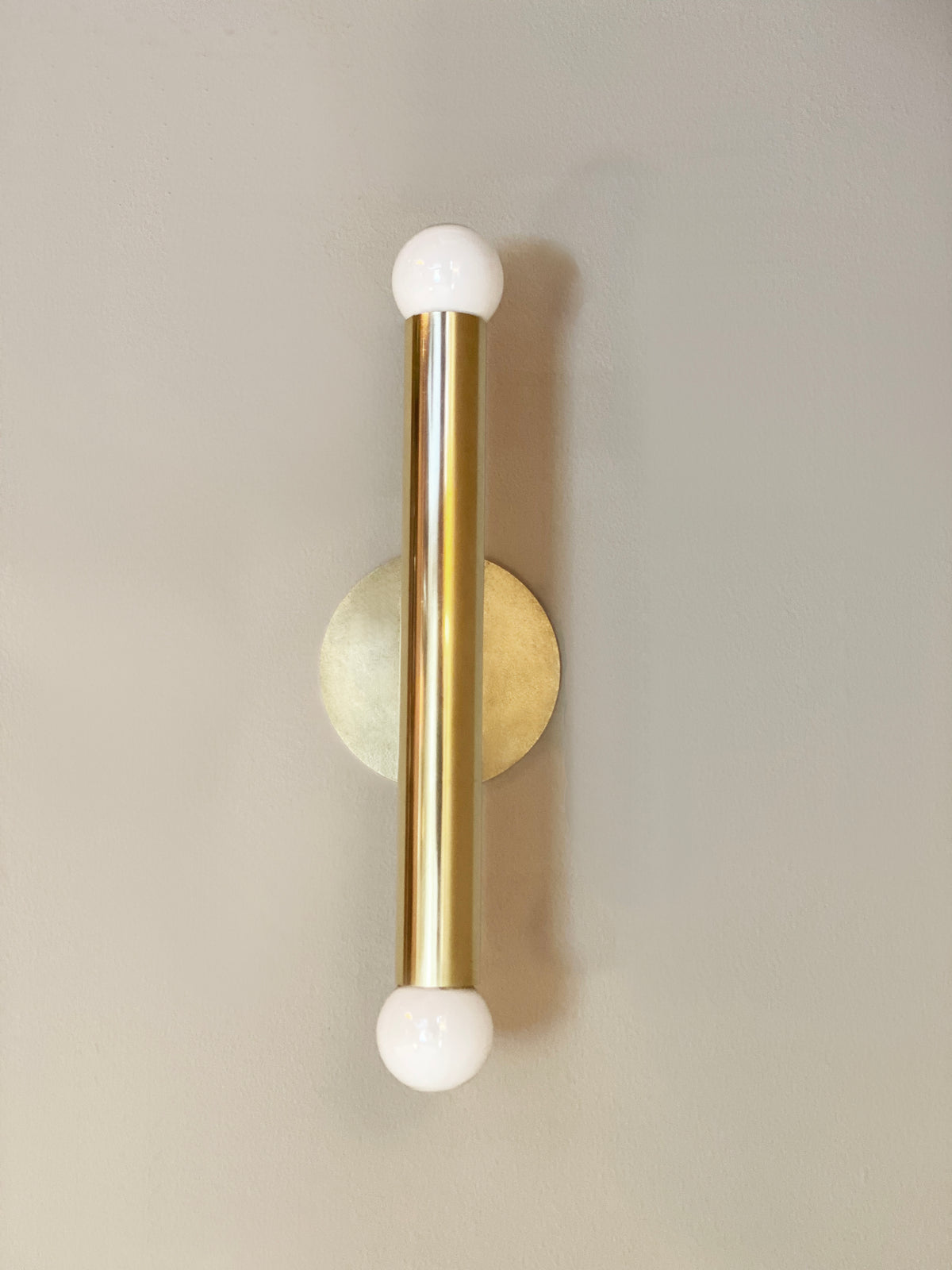 The Miro 2 - 12" Sconce by Long Made Co. is a modern handmade brass wall fixture with a vertical cylindrical design and round white bulbs on both ends, ideal for elegant vanity lighting.