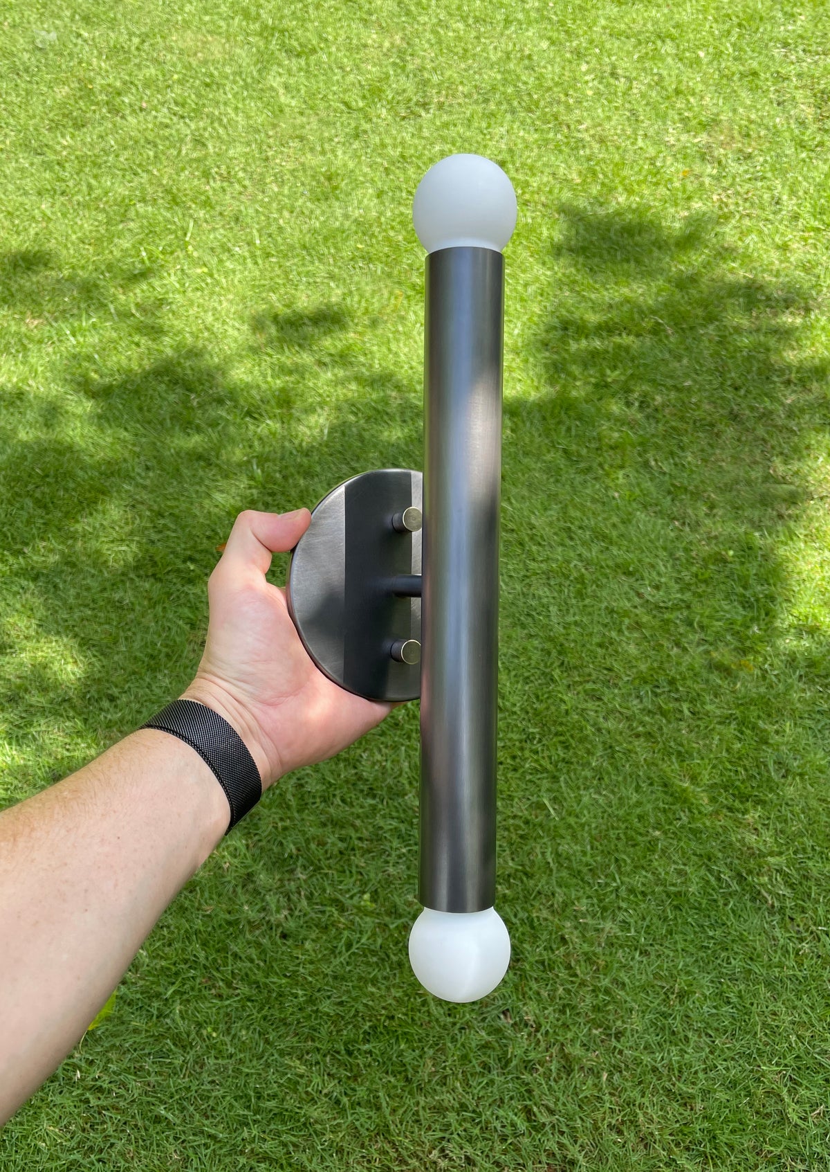 A person holds the Long Made Co. Miro 2 - 12" Sconce, featuring a modern metal cylindrical design with round white ends and a circular brass mounting plate, adding sleek style to any space over a grassy lawn.