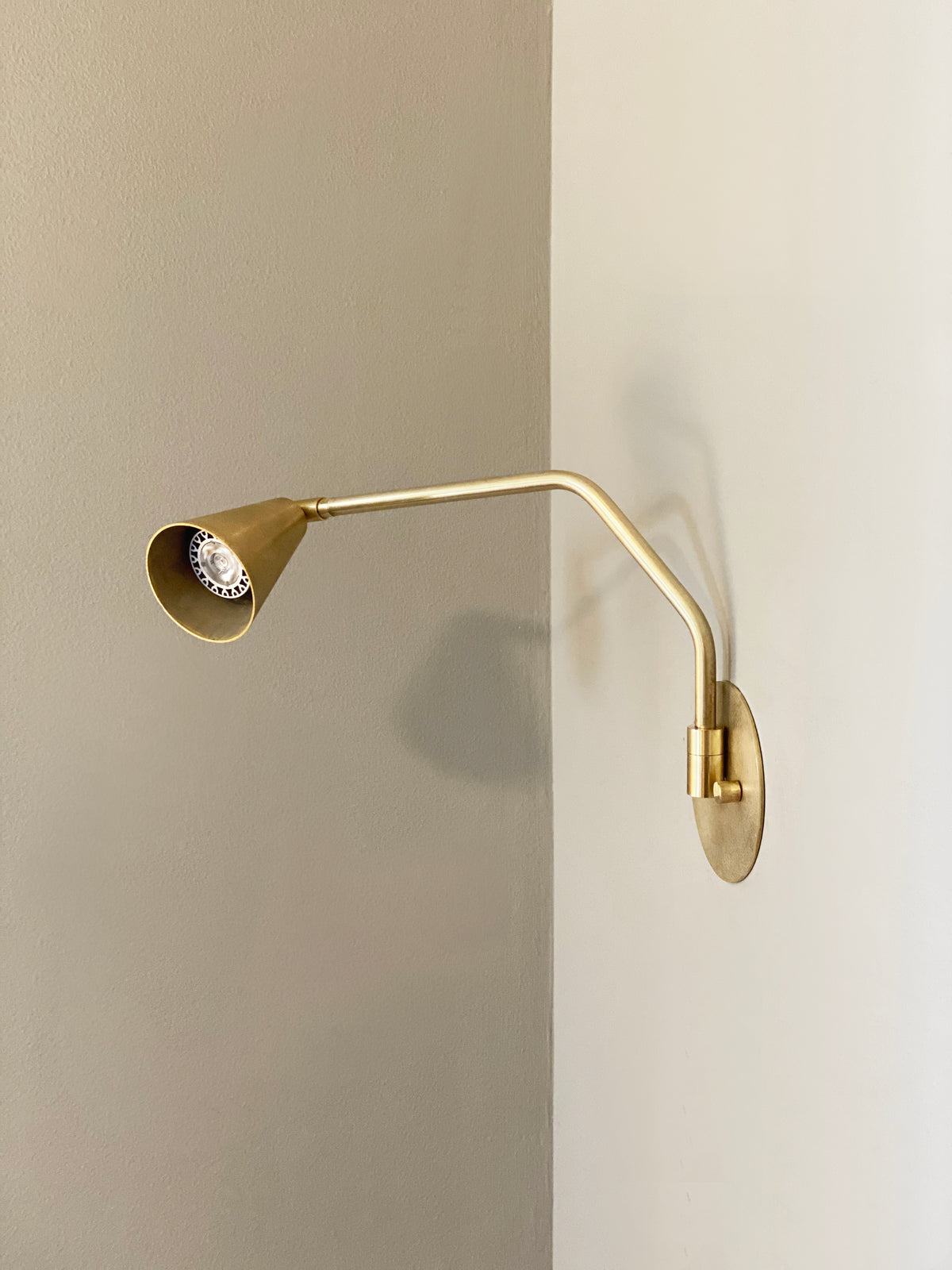 The Tiel Arch Wall Lamp by Long Made Co., featuring an angular brass arm and conical shade, is mounted on a two-tone wall and casts a faint shadow.