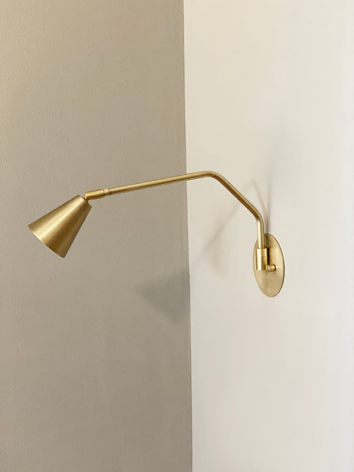 The Tiel Arch Wall Lamp by Long Made Co. is a modern handmade brass fixture with a conical shade and adjustable arm, hardwired to a white wall and extending over a corner with a neutral background.