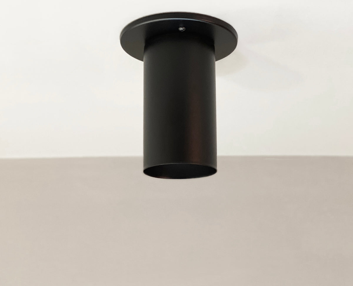 A black solid steel Cove Flush Spot by Long Made Co. from the Cove Series is mounted on a white ceiling above a light gray wall, creating a sleek, minimalist look.