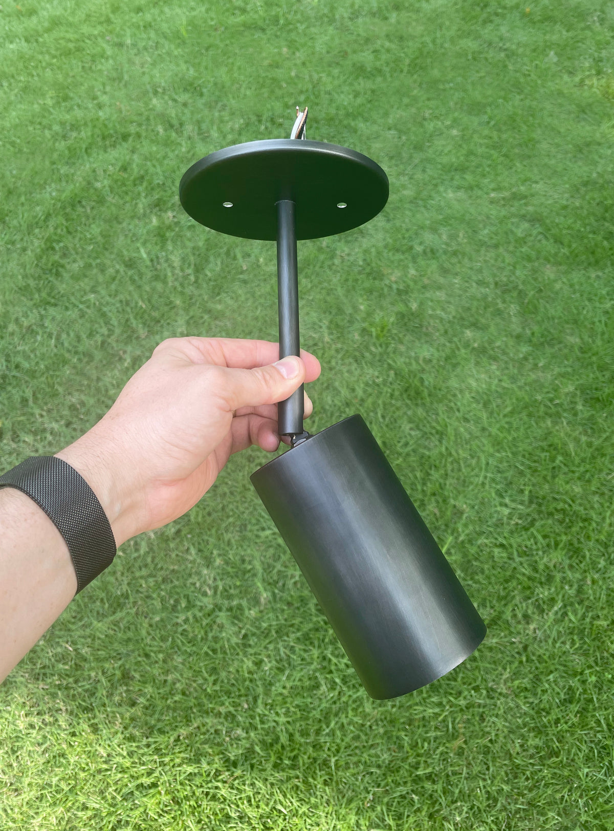 A hand holds the Long Made Co. Cove Drop Spot, a black minimalist outdoor spotlight with a round mounting plate, set against green grass.