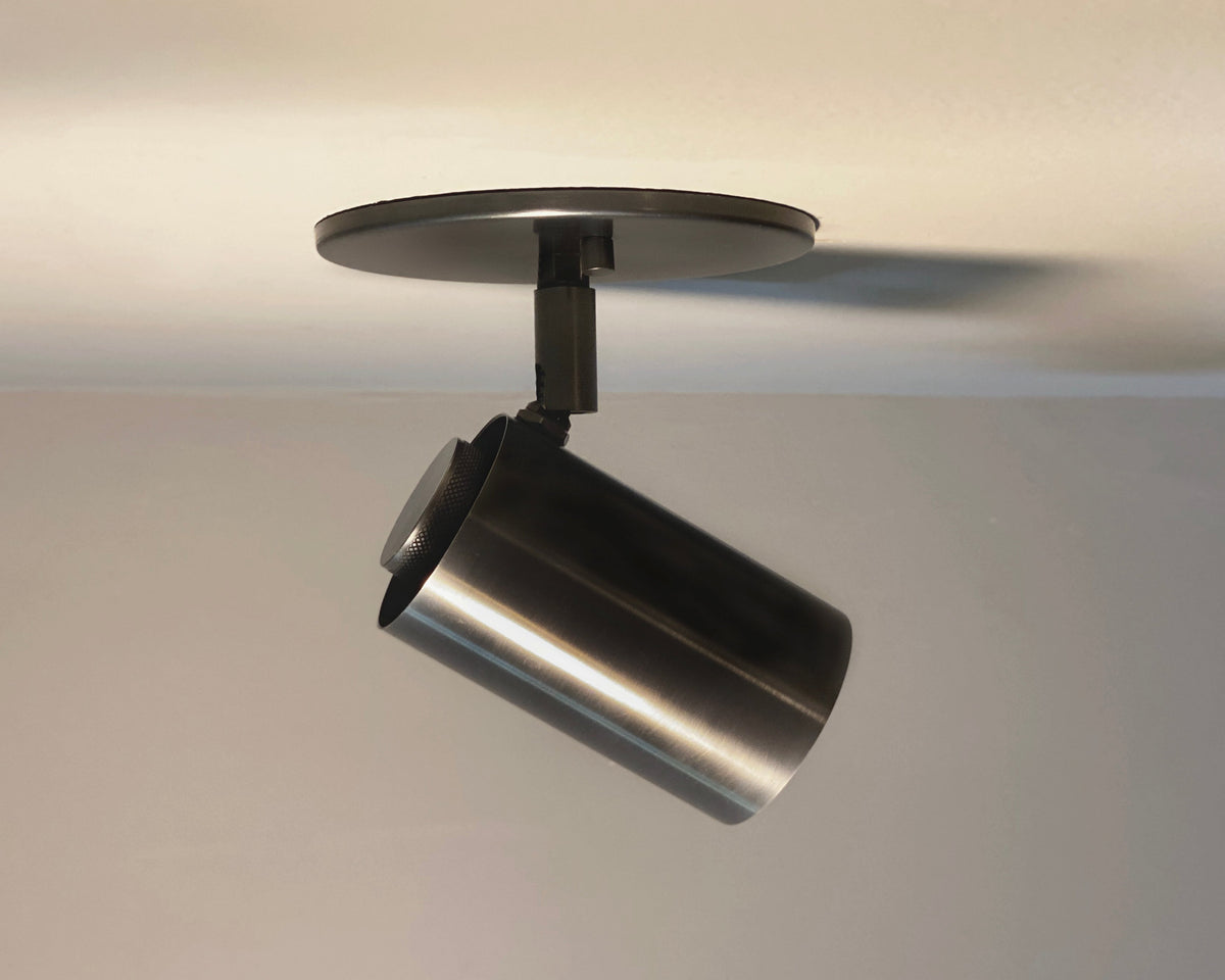 The Brass Cylinder Adjustable Spot - Side Mount by Long Made Co. is a modern ceiling spotlight with a cylindrical brass body, adjustable head, and round base, designed for side mounting on a white ceiling.