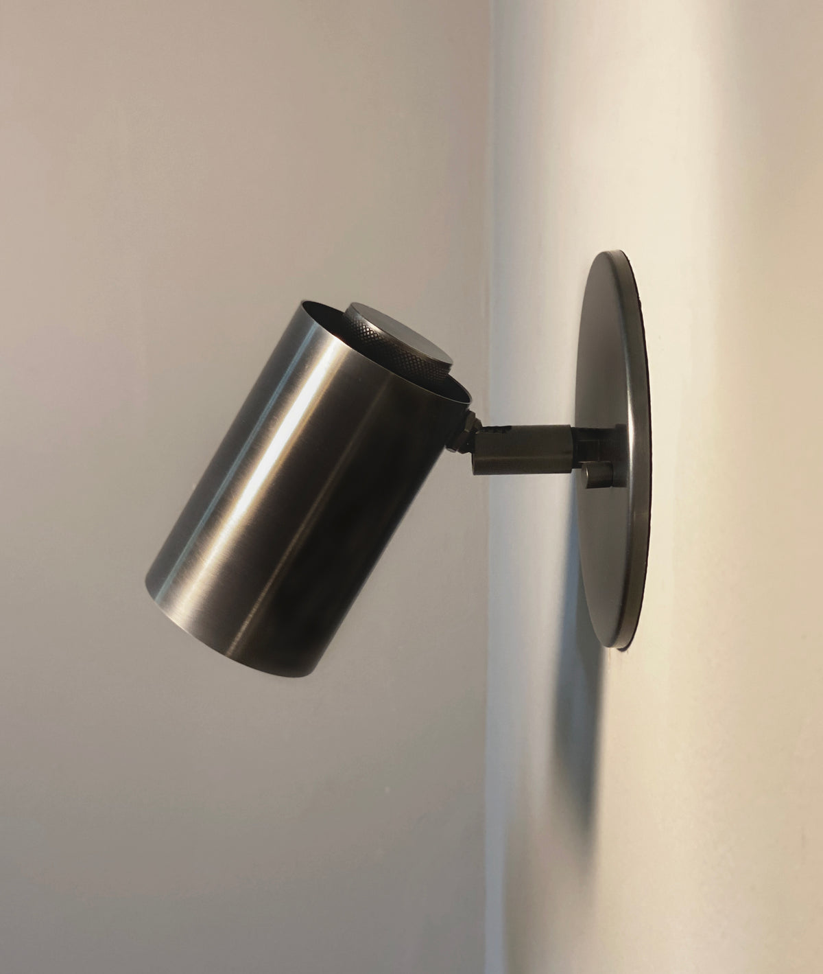 The Brass Cylinder Wall Lamp by Long Made Co. features a modern cylindrical brass design with a round base, mounted on a white wall and angled downward, combining classic style with mid-century modern functionality.