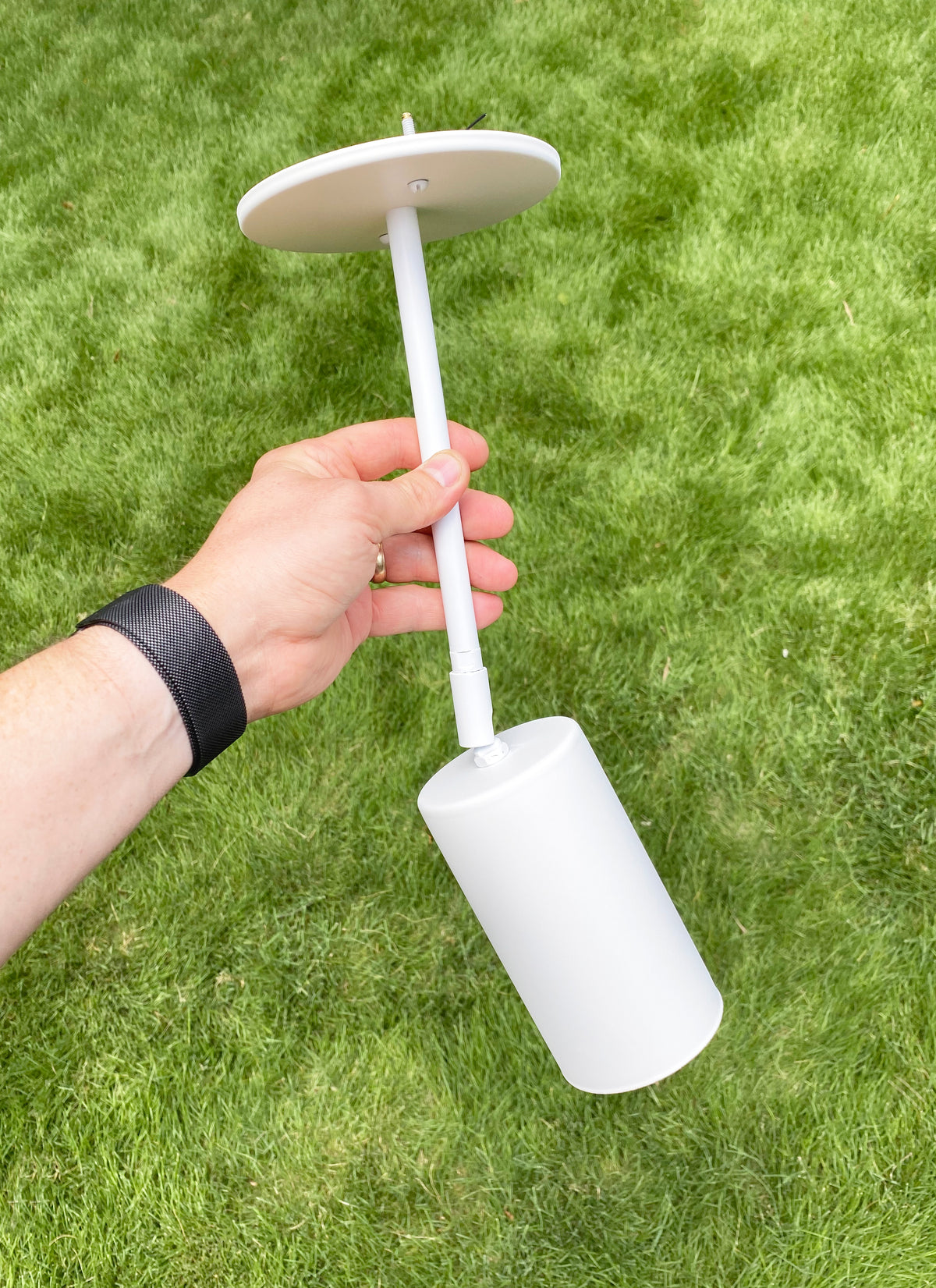 A hand holds the Cove Drop Spot by Long Made Co.—a minimalist white cylindrical spotlight attached by a rod to a flat, round disc—set against green grass.