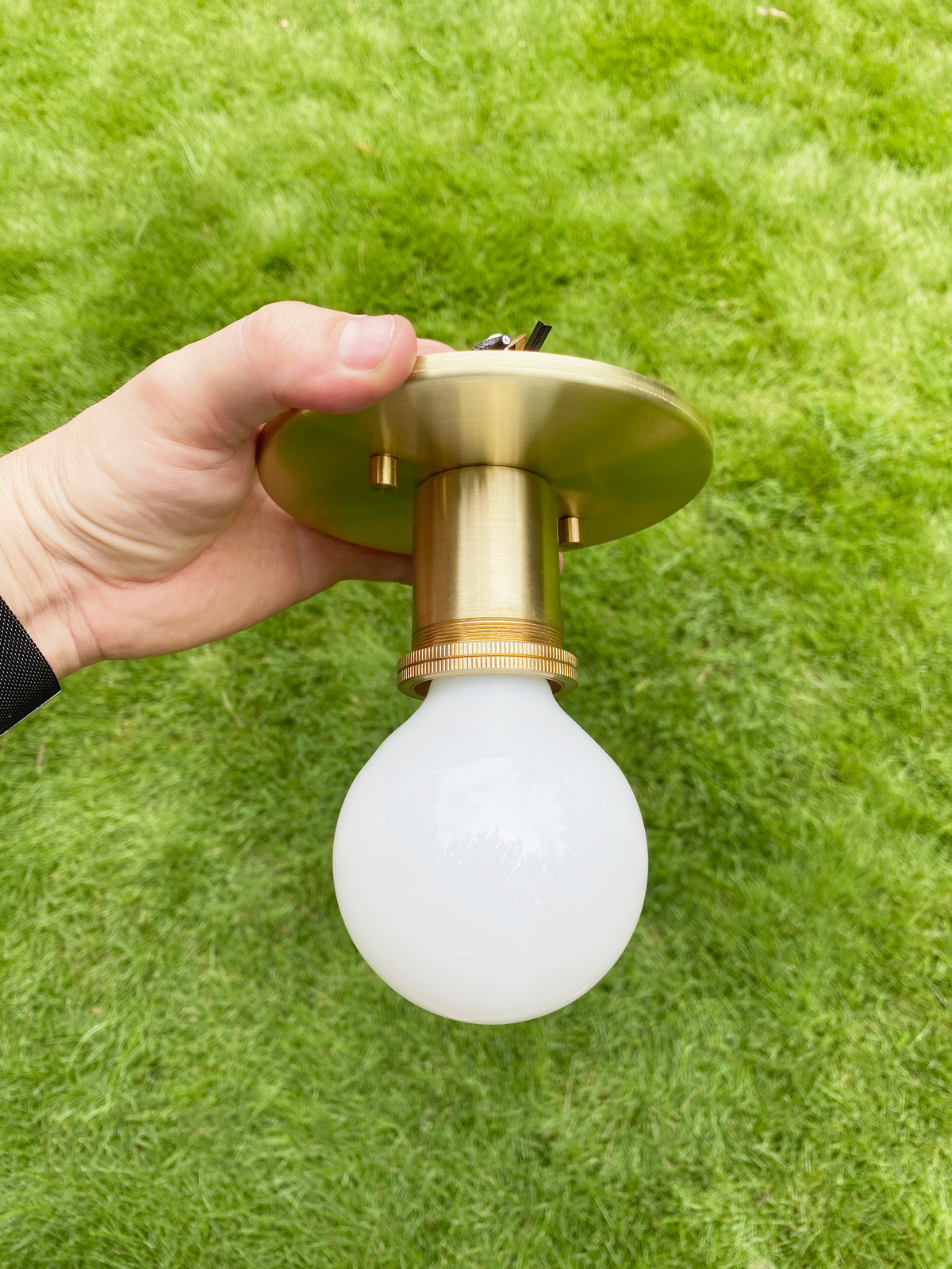 A hand holds the Brass Minimal Flush by Long Made Co., featuring a round white bulb, set against green grass.