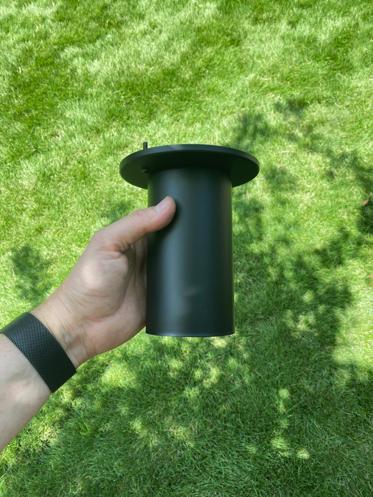 A person with a black wristband holds the Long Made Co. Cove Flush Spot, a solid steel black cylindrical downlight with a flange, while standing on green grass in bright sunlight.
