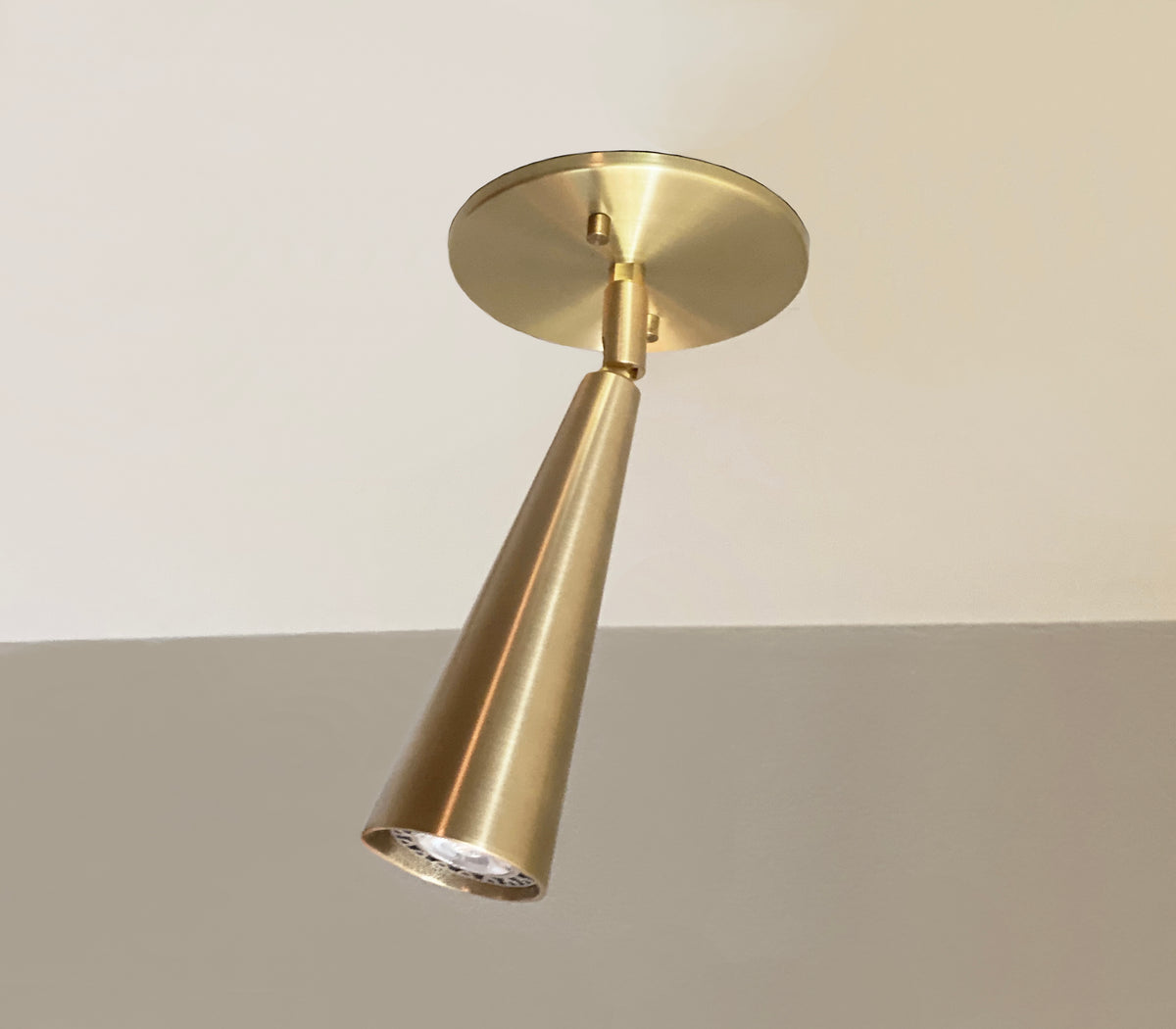 The Pine Adjustable Spot by Long Made Co. is a handmade brass cone-shaped directional ceiling downlight, mounted on a white and beige ceiling and viewed from below.