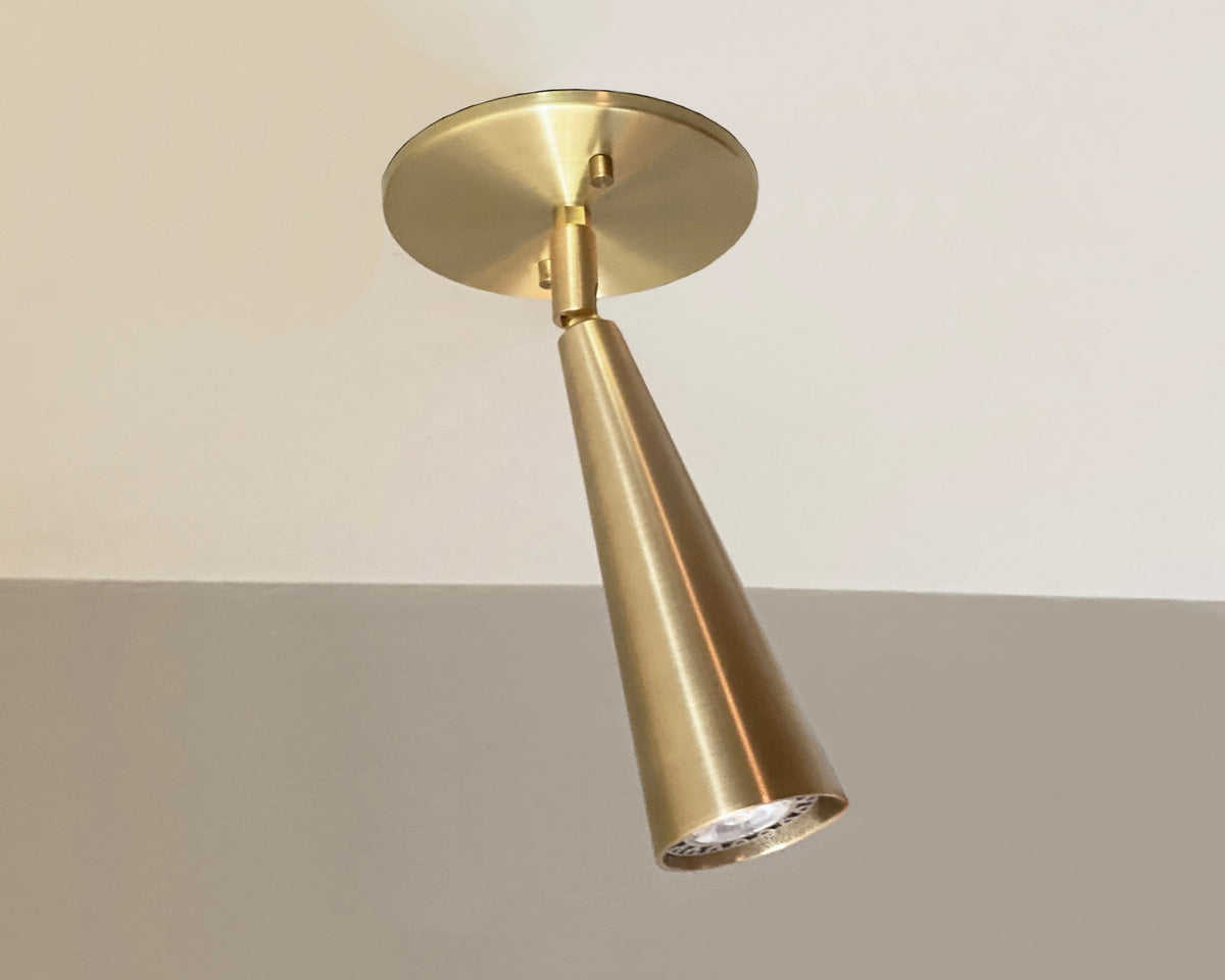 The Pine Adjustable Spot by Long Made Co. is a modern handmade brass cone-shaped spotlight suspended from a circular mounting plate on a white ceiling.