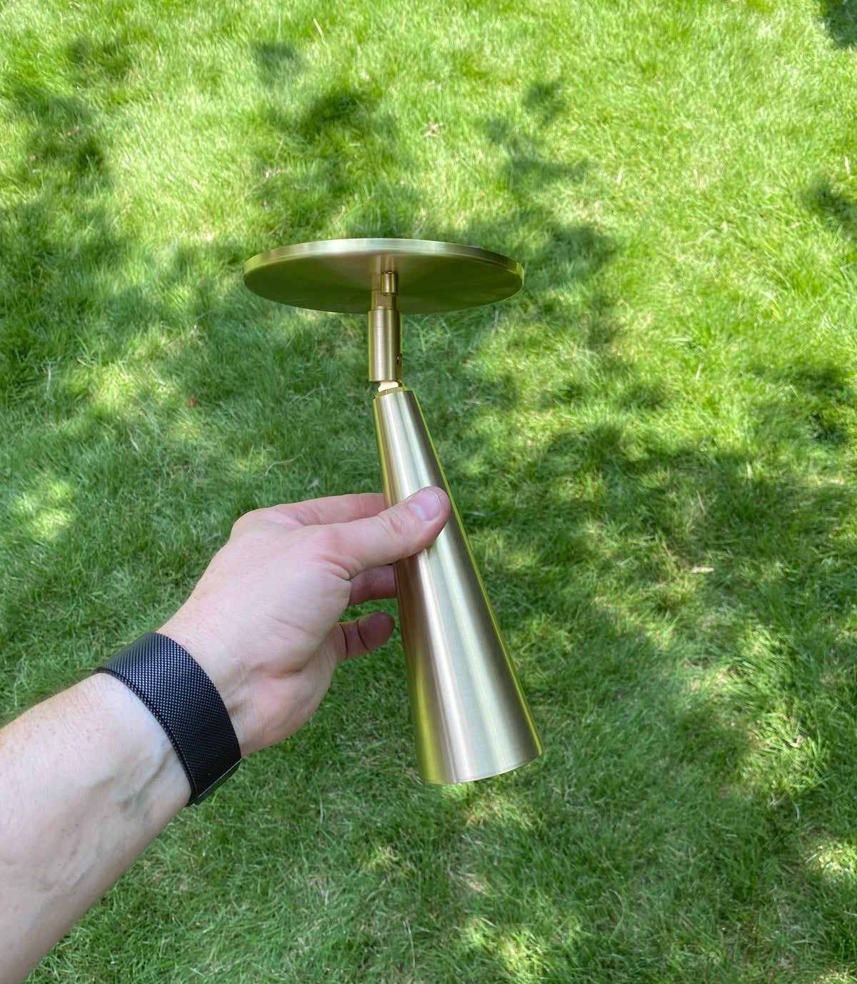A person wearing a black wristband holds the Long Made Co. Pine Adjustable Spot, a handmade brass cone-shaped ceiling light, outdoors over a green grassy lawn.