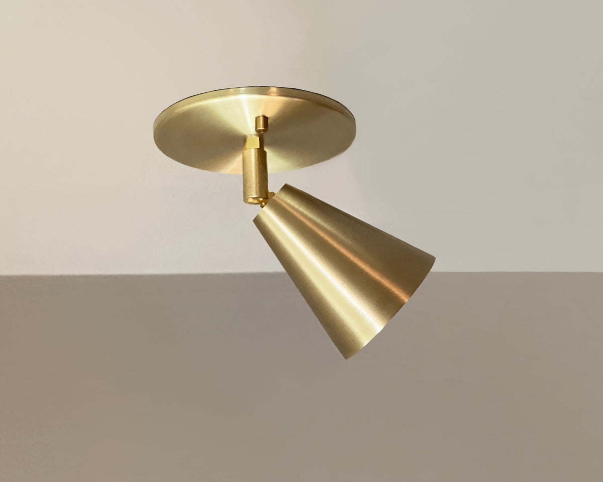 The Tiel Spot - Adjustable by Long Made Co. is a sleek, modern solid brass ceiling light with a conical shade, offering an elegant alternative to recessed can lights when mounted on a white and light gray ceiling.