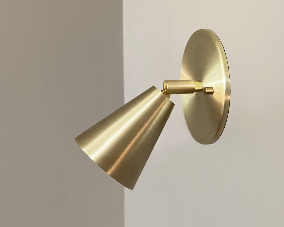 The Tiel Wall Spot by Long Made Co. is a handmade brass wall sconce with a conical shade and circular base, offering a minimalist, sleek design perfect for light-colored walls.