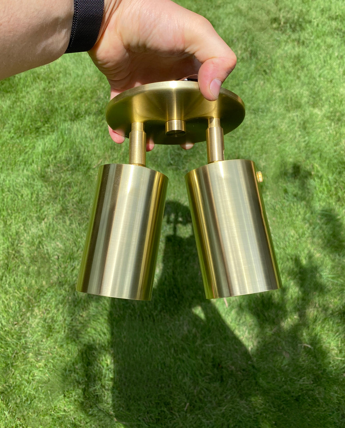 A hand holds the Long Made Co. Brass Cylinder Double / Triple Spot, an adjustable brass-colored fixture, outdoors with green grass in the background.