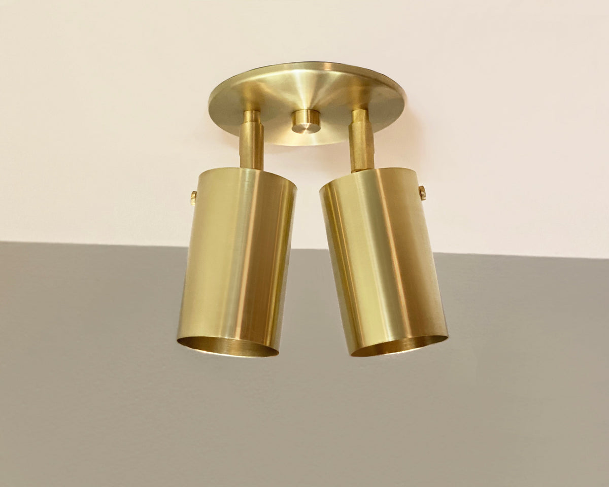 Double brass cylinder spotlights mounted on a ceiling, with adjustable heads positioned vertically and horizontally.