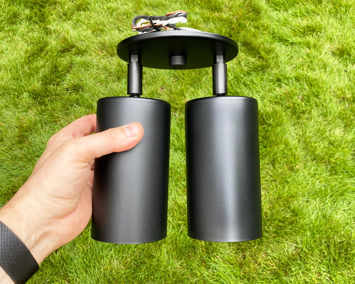 A hand holds the Long Made Co. Cove Double / Triple Spot, a black outdoor light fixture with solid steel spotlights and exposed wires, set against green grass.