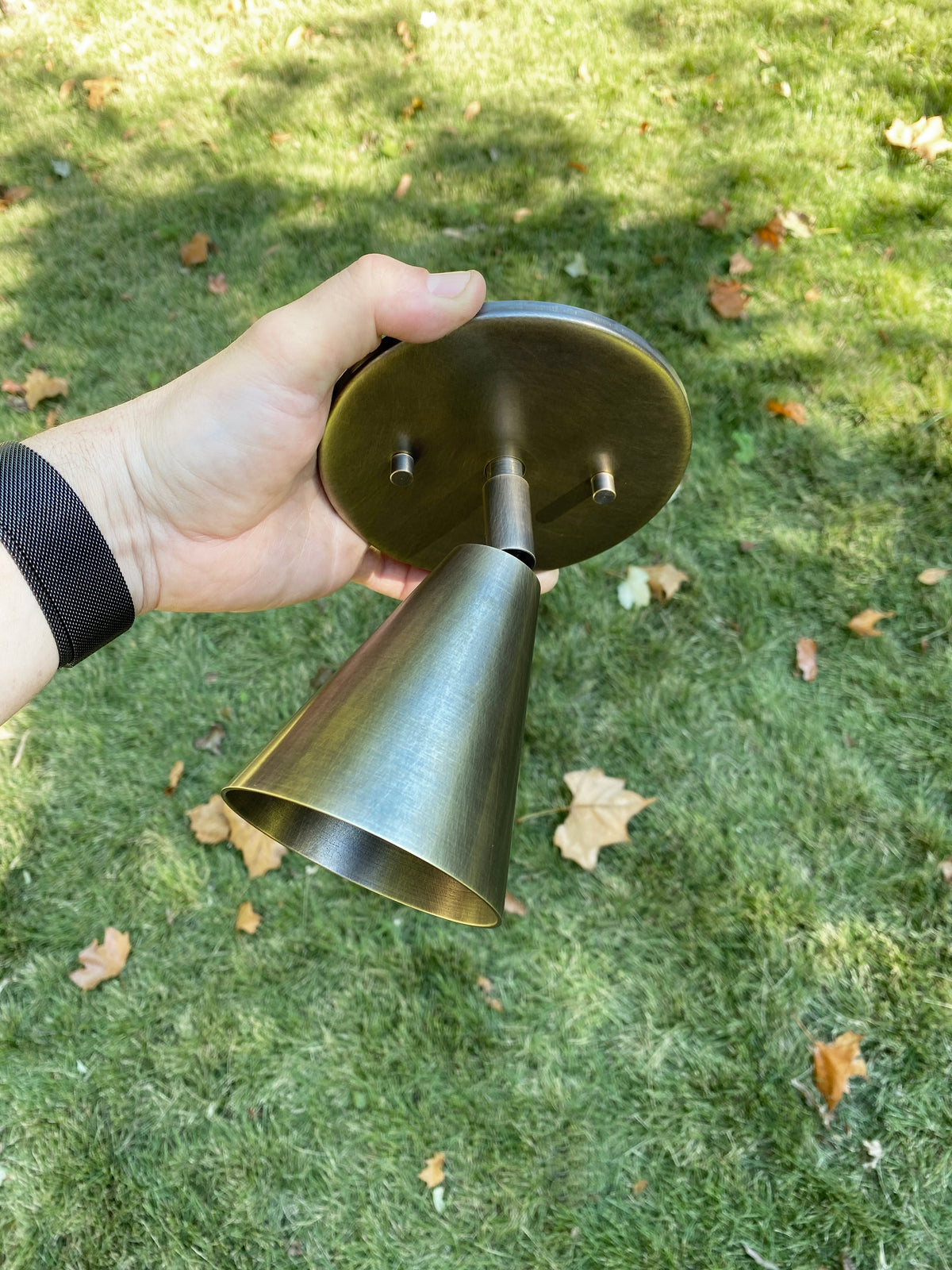 A hand displays the Tiel Wall Spot by Long Made Co., a brass spotlight ceiling wall fixture, outdoors over green grass with brown leaves, highlighting its handmade lighting style.