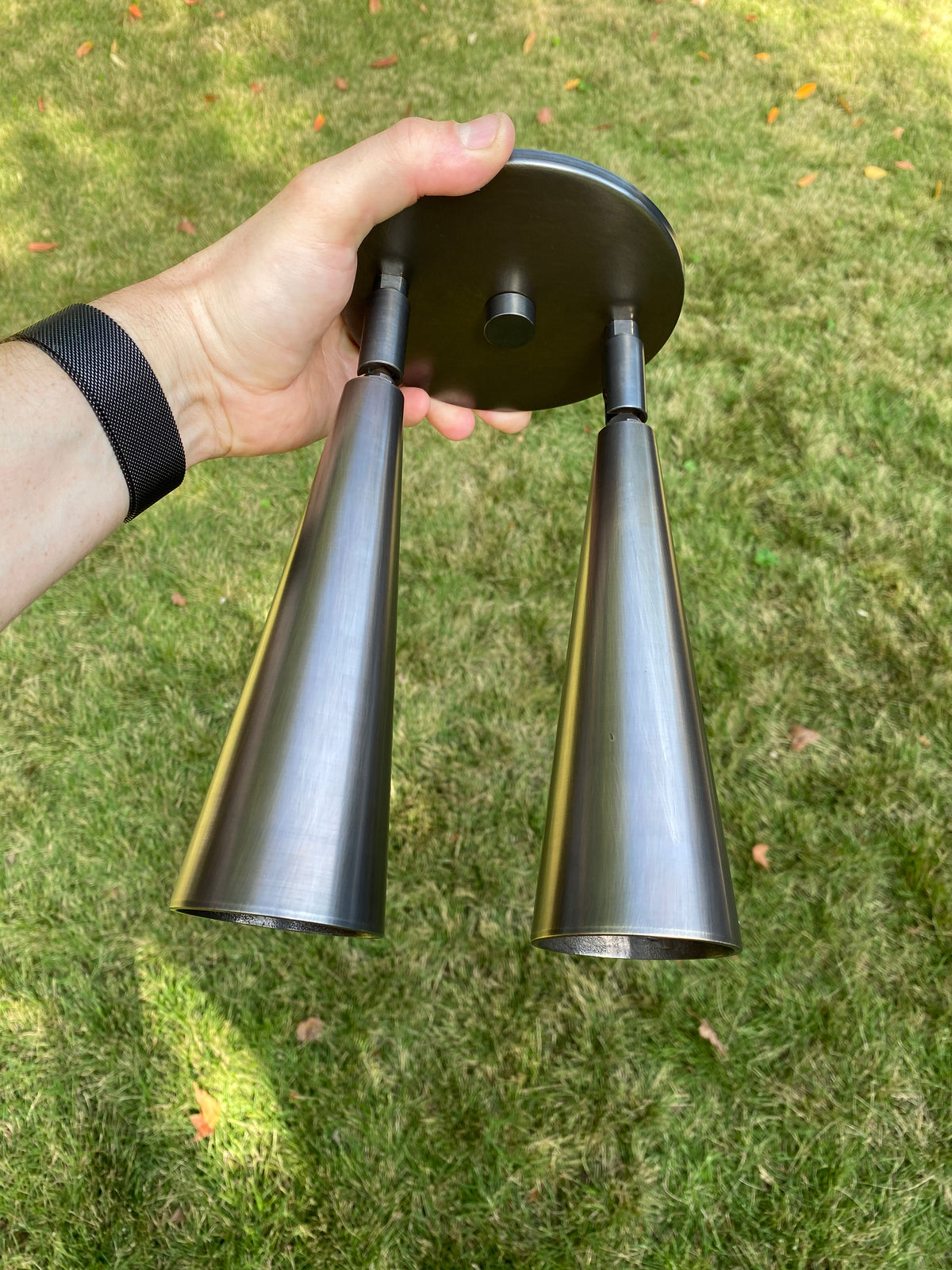 A hand holds the Long Made Co. Pine Double / Triple Spot, a metallic fixture with adjustable spot heads, outdoors against green grass.